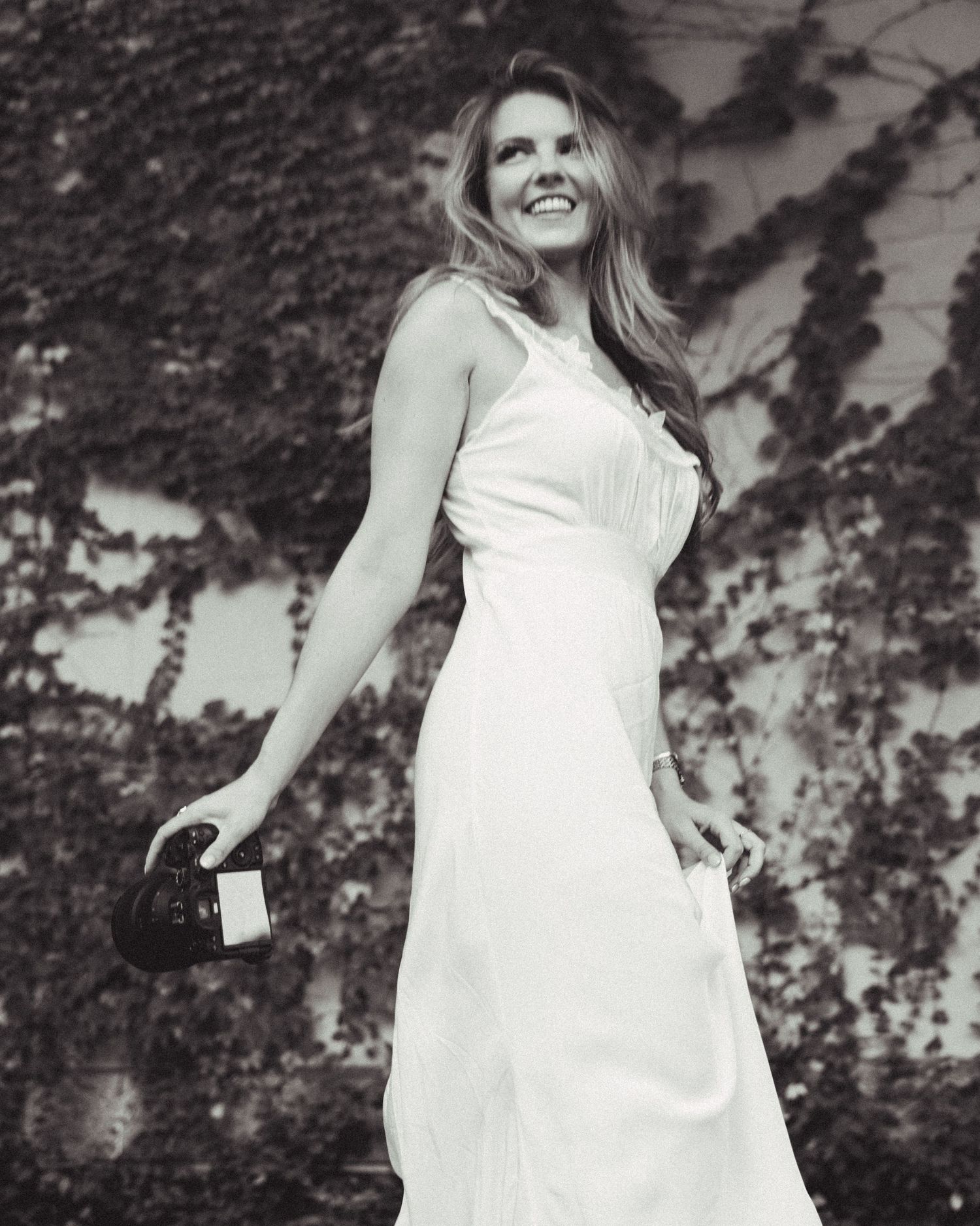 Black and white photo of a smiling woman in a white dress holding a purse, standing in front of ivy-covered wall.
