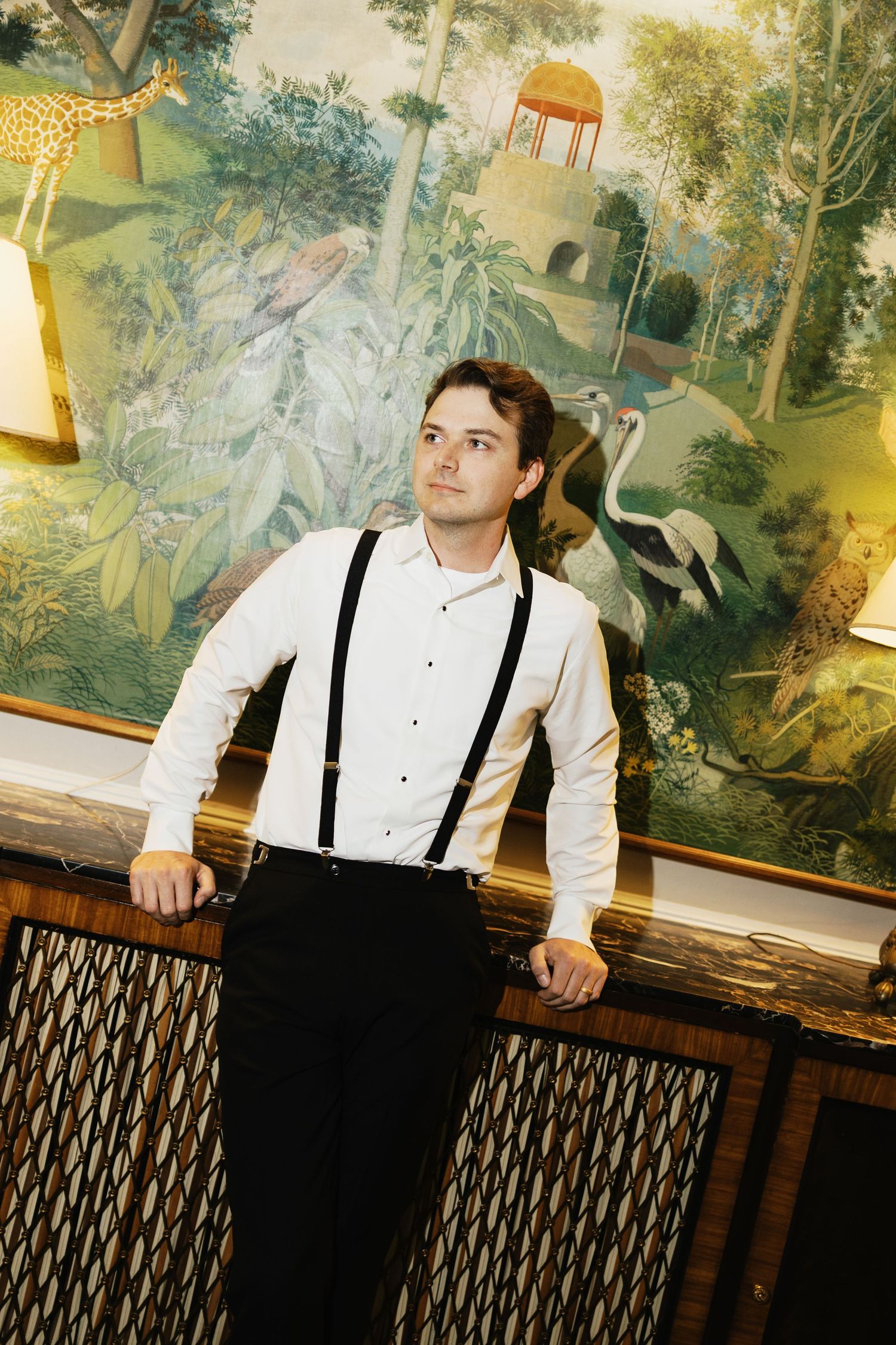 Young man in white shirt and black suspenders leaning on railing before vibrant jungle mural.