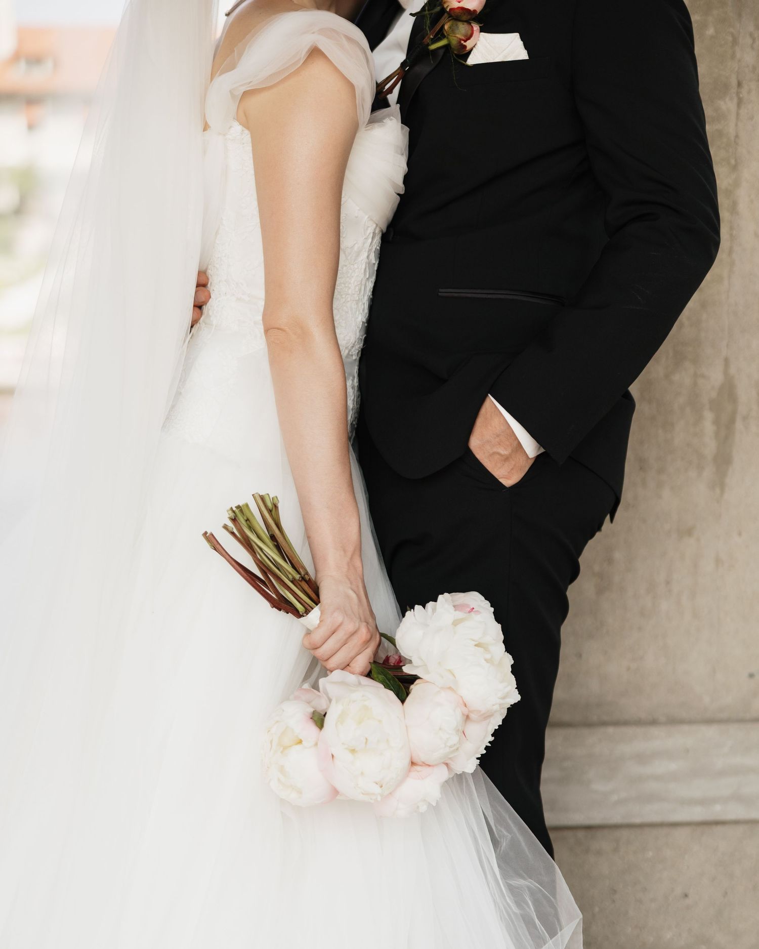 Bride holding blush peony bouquet in white tulle gown beside groom in black suit near stone column.