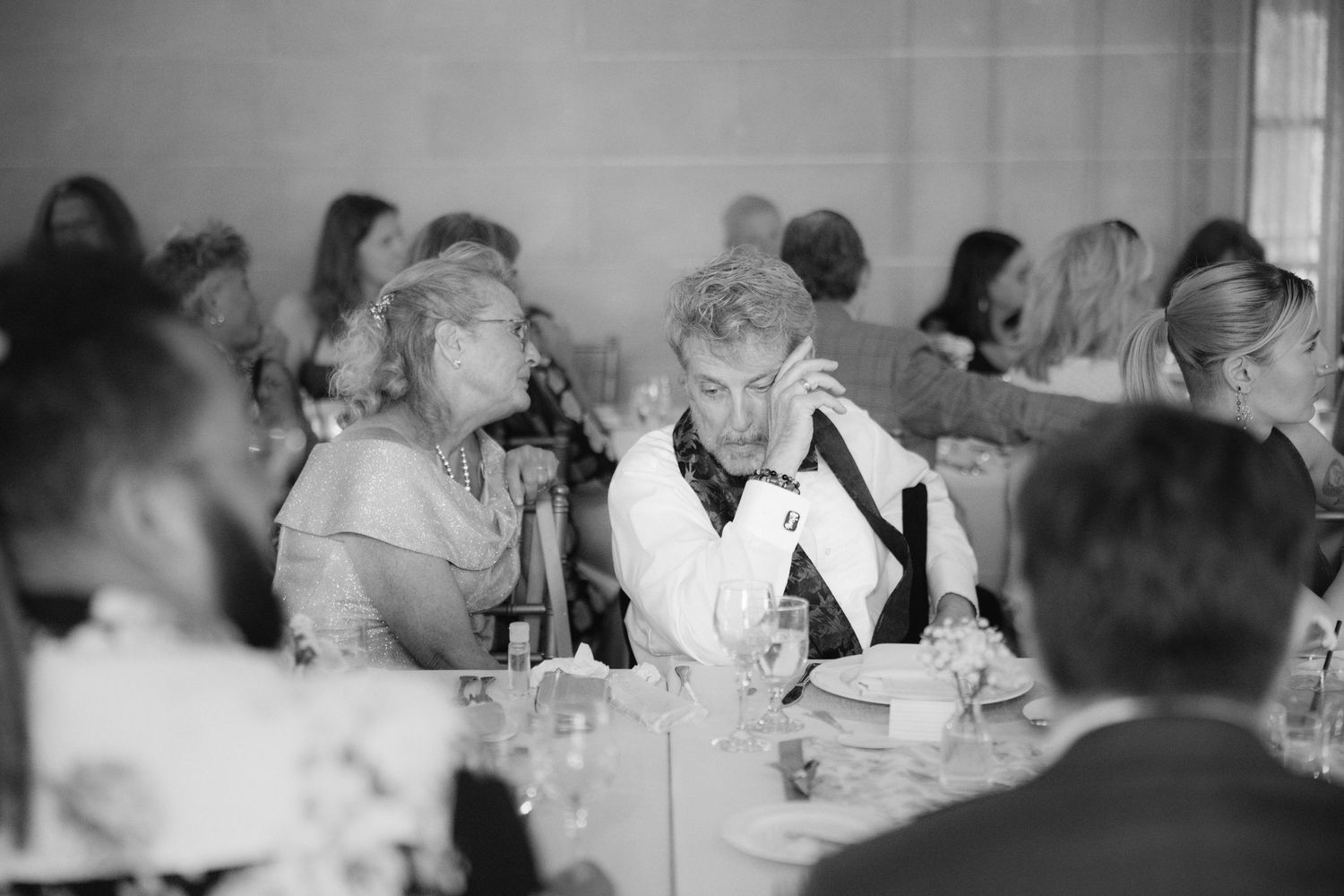 Black and white wedding reception photo of guests seated at tables, engaged in conversation in an elegant venue.