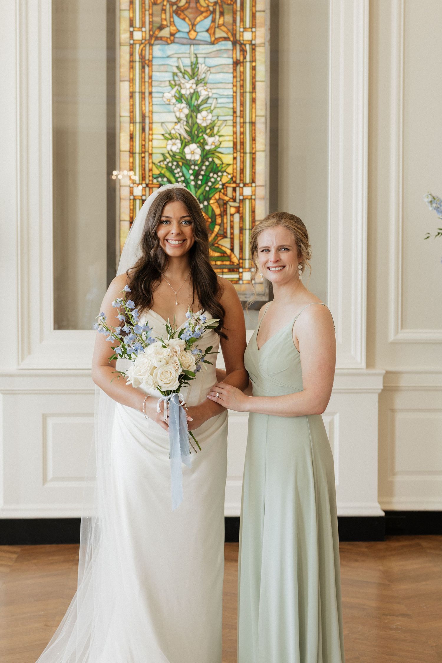 Wedding party in sage green bridesmaid dresses and white bridal gown pose in elegant venue with ornate gold decor.