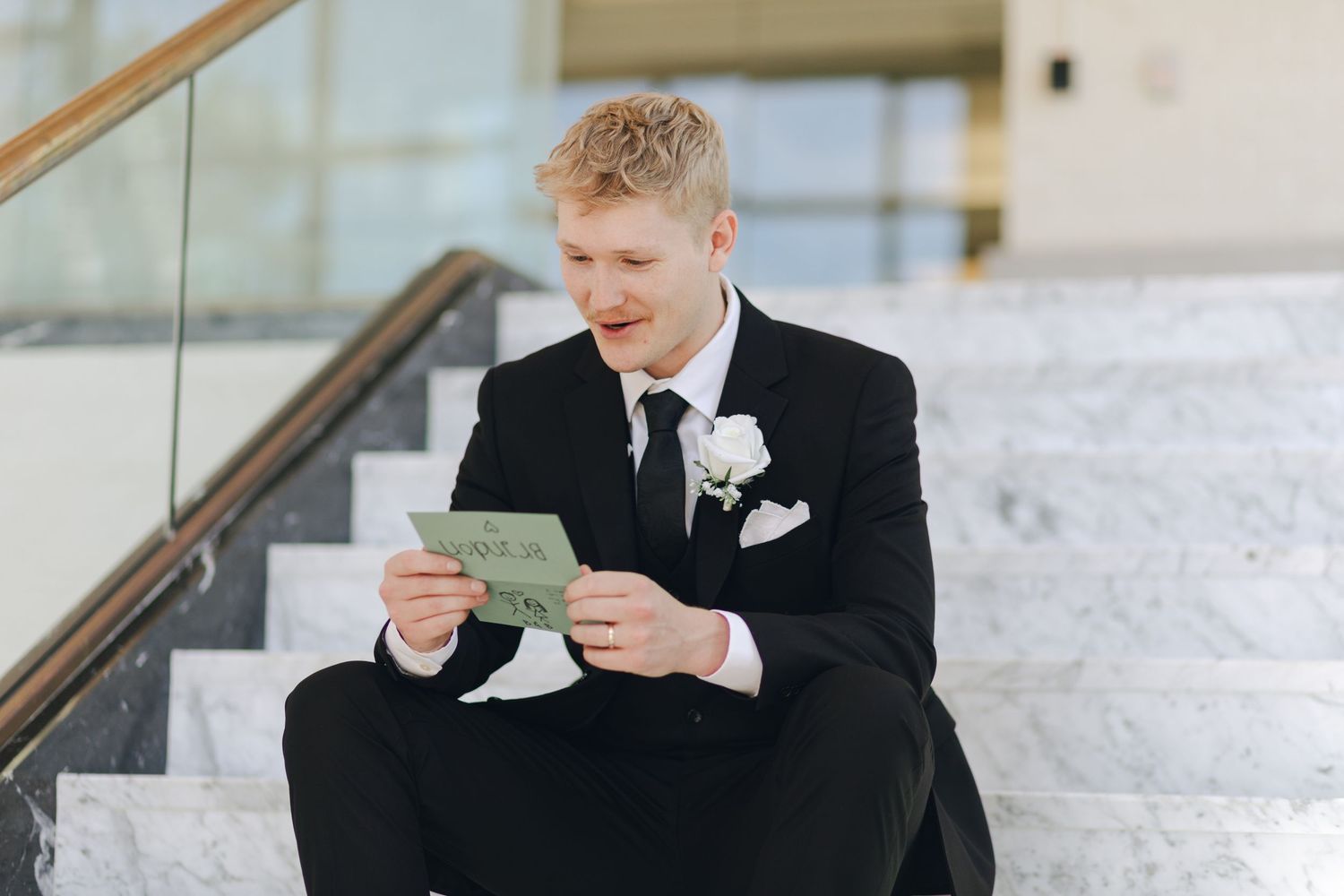 A person in a black suit holds and reads from a paper while sitting on marble steps.