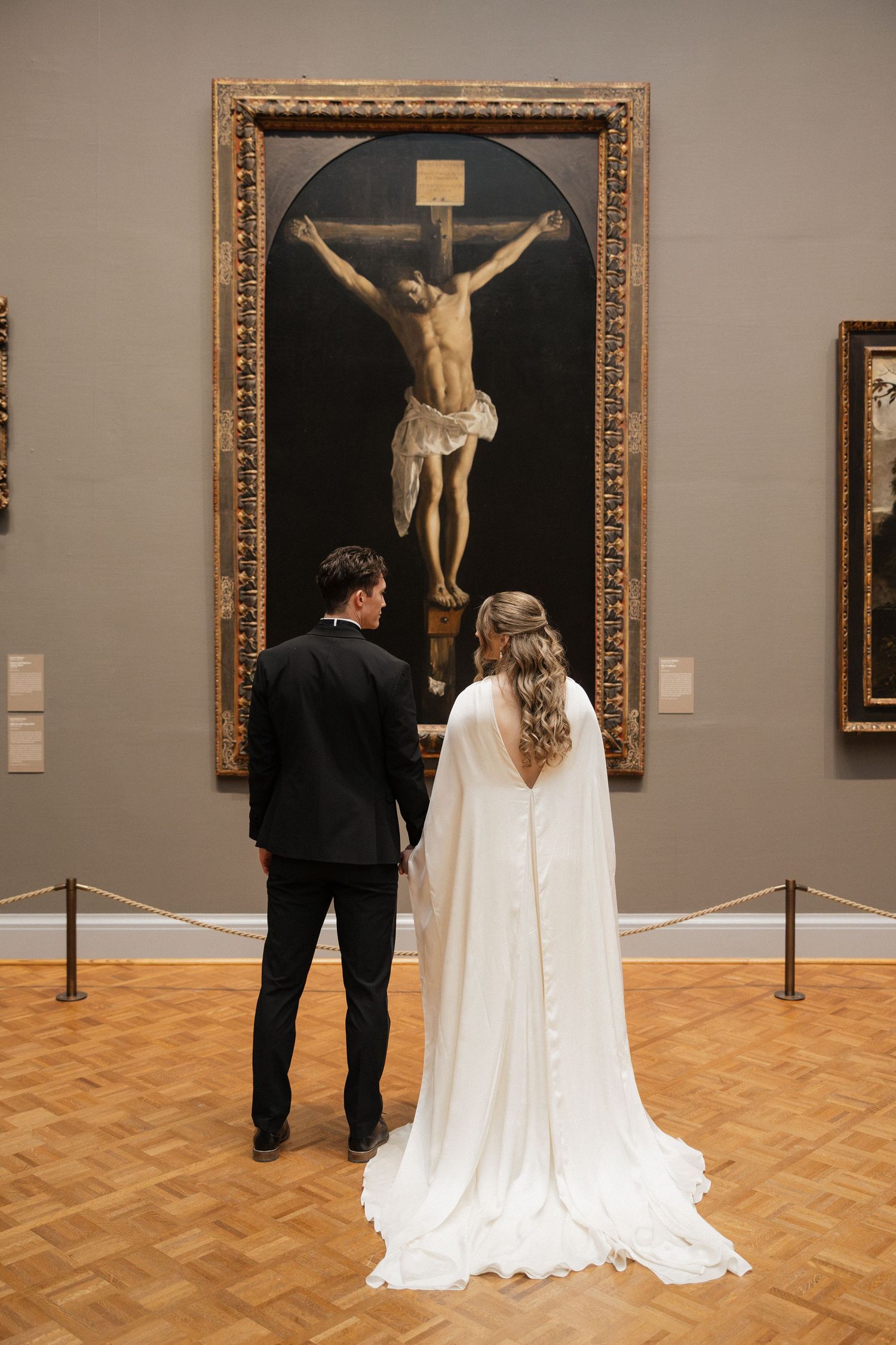 A couple in wedding attire views religious artwork featuring crucifixes in an art gallery with wooden floors.