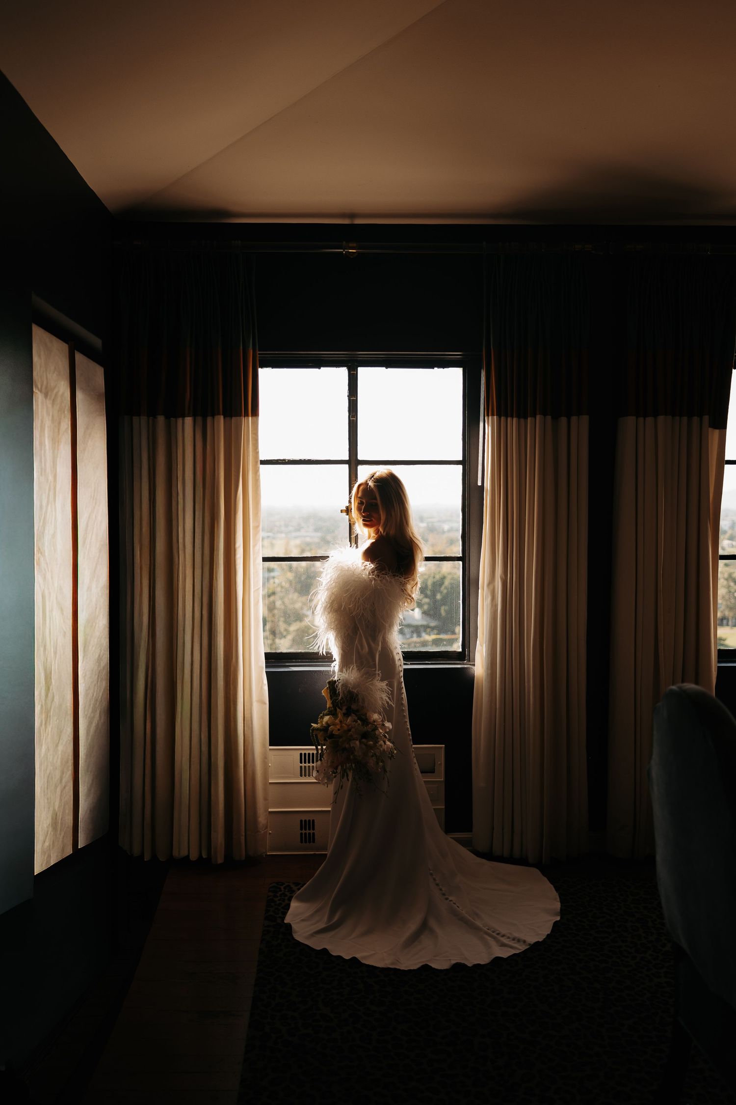 A silhouetted figure in a wedding dress stands by a window in a dark hotel room creating a dramatic moody atmosphere.