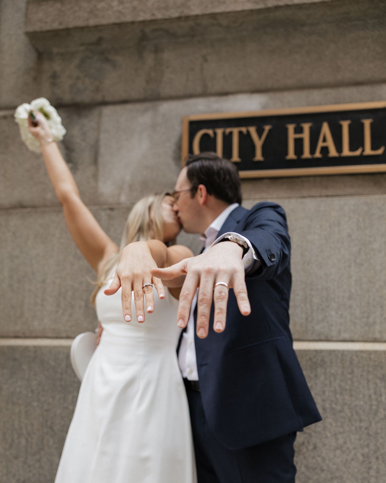 Newlyweds kissing outside City Hall, showing off wedding rings, bride holding bouquet aloft in white dress and groom in navy suit.