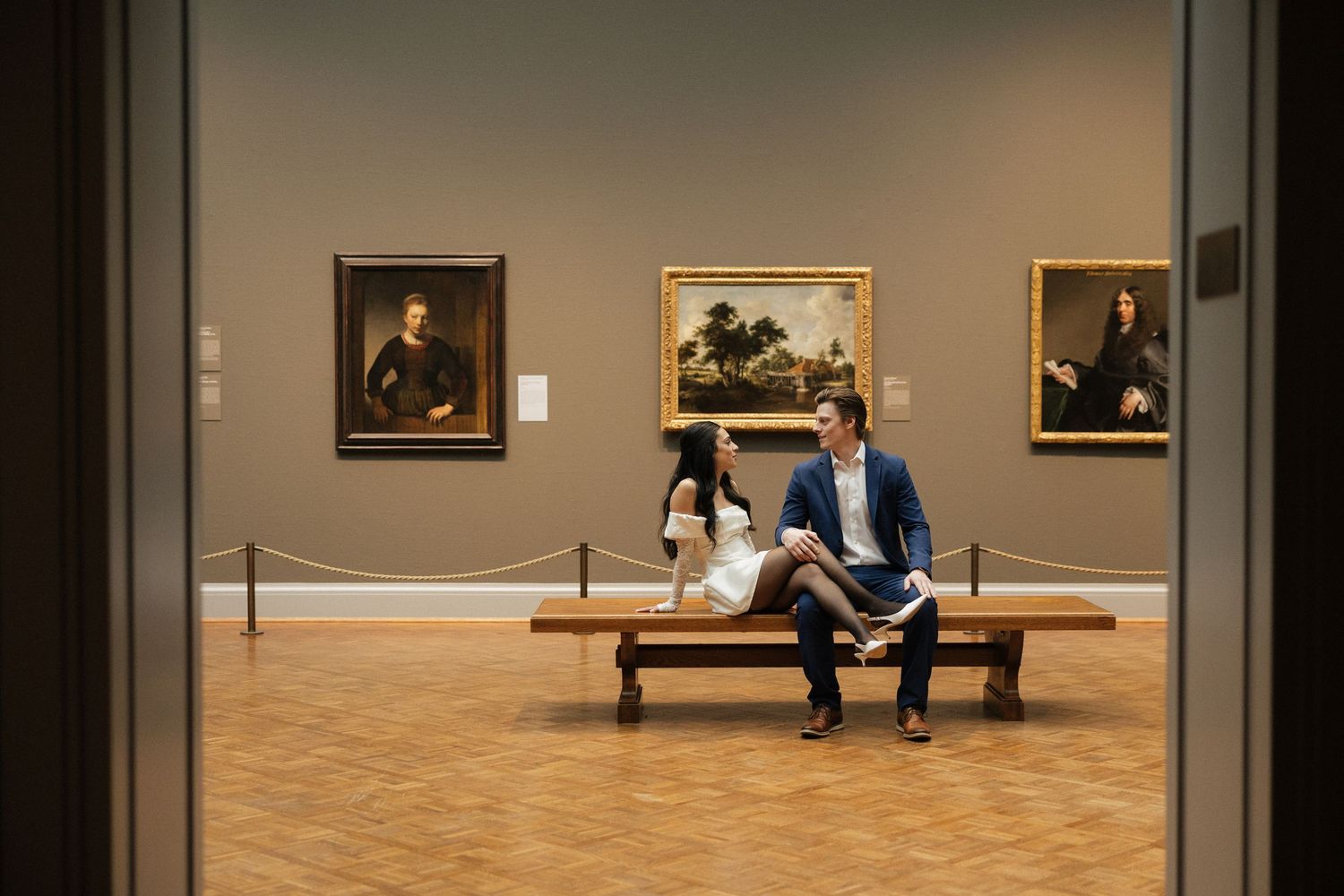 Two visitors sit on a wooden bench viewing classical portrait paintings in an art museum gallery.