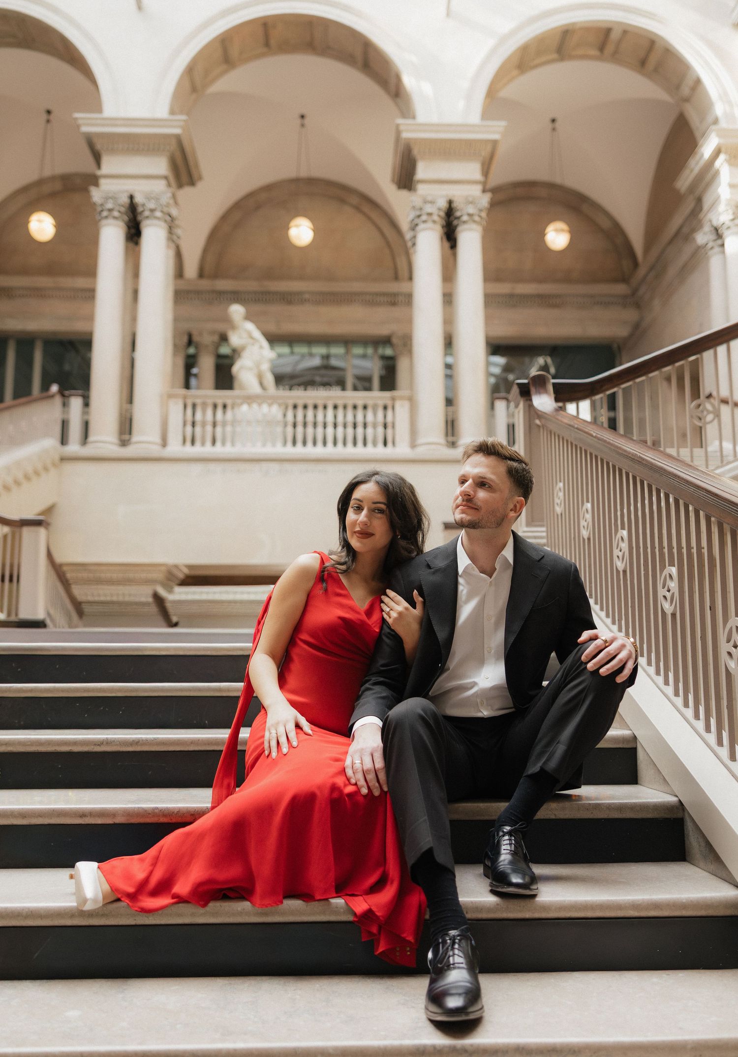 A couple in formal attire poses together on marble steps in an elegant indoor venue with arched columns and warm lighting.