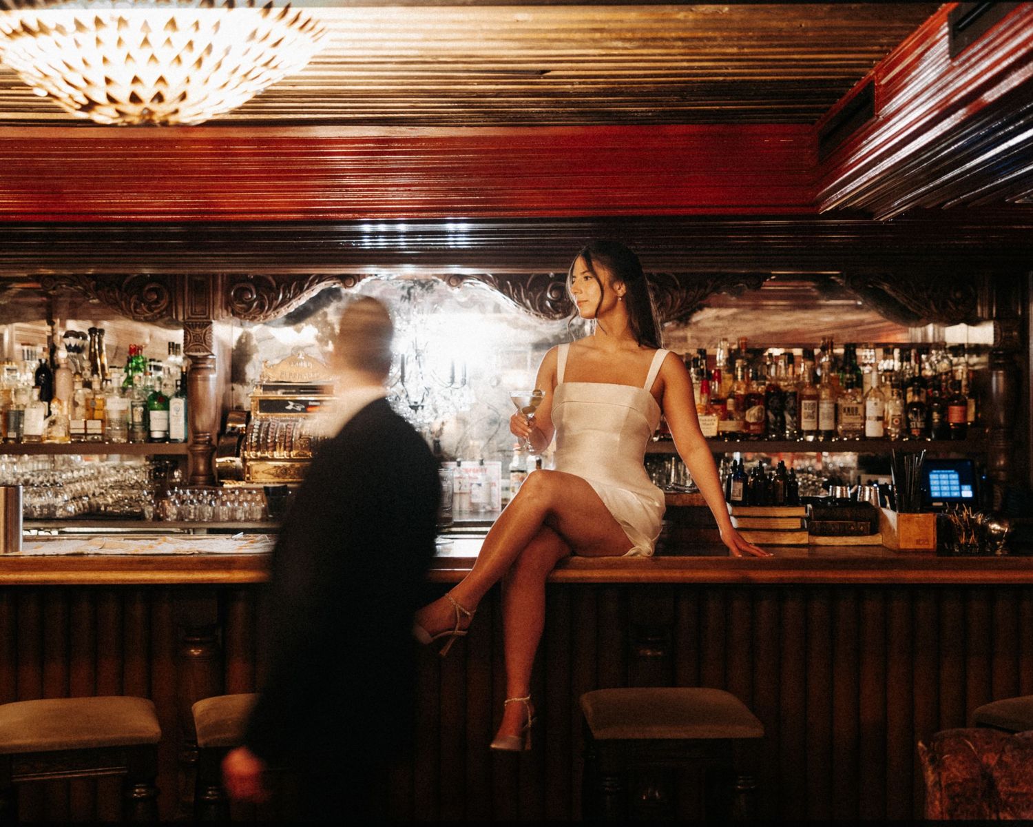 People gather at a dimly lit bar counter with warm ambient lighting and bottles displayed on shelves.