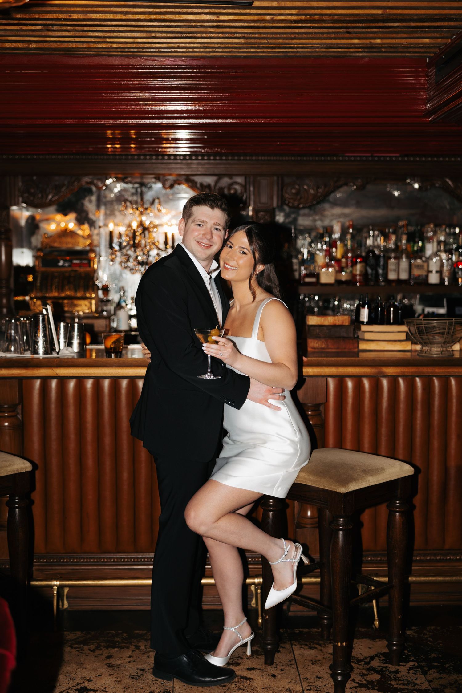 Couple in wedding attire embraces playfully at an elegant bar with wood paneling.
