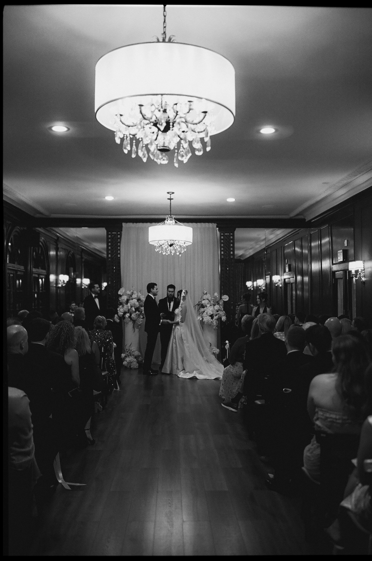 Black and white photo of elegant indoor wedding ceremony under crystal chandelier with seated guests.