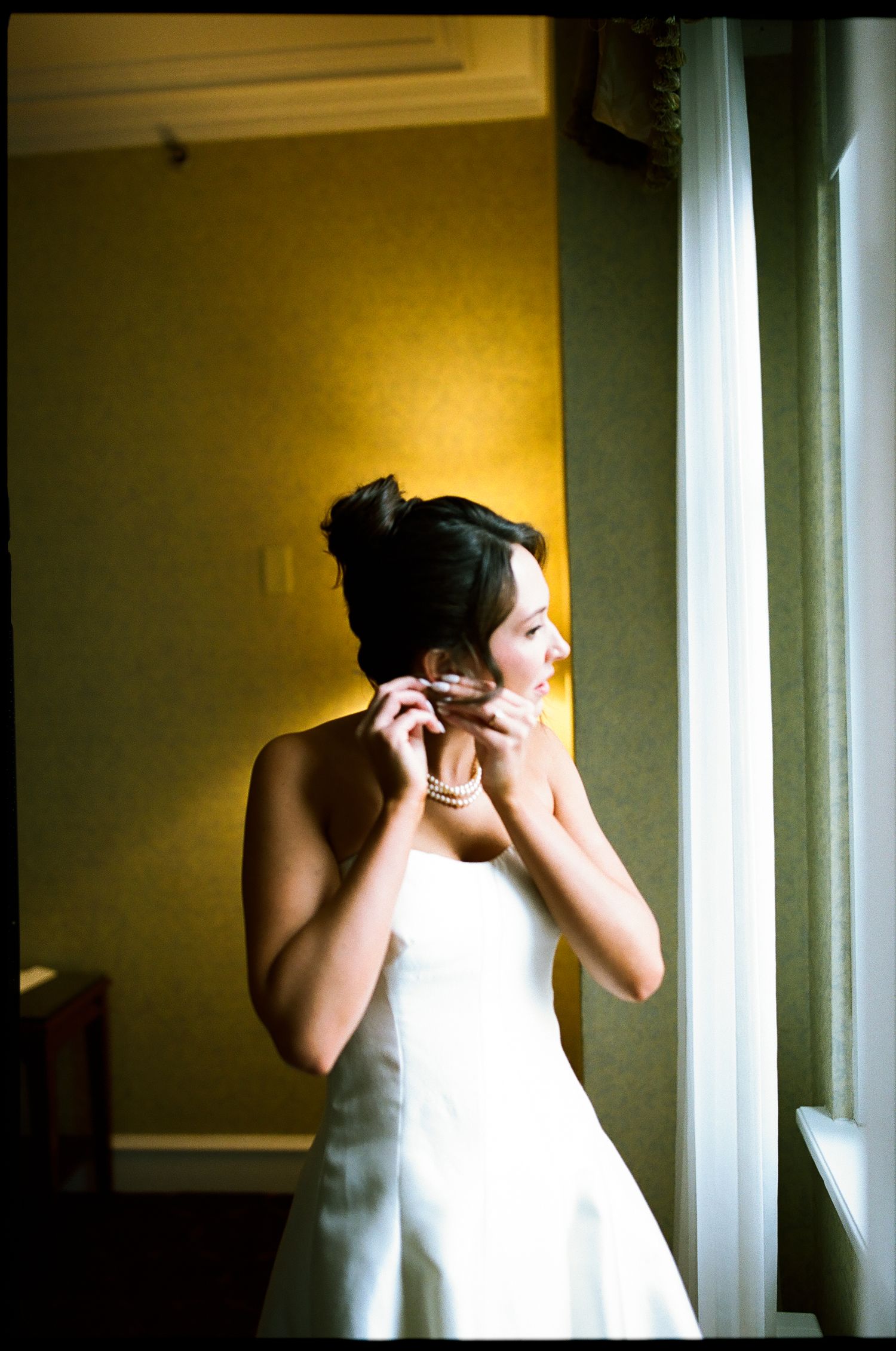 Bride in white strapless gown putting on earrings by window in golden-lit hotel room.