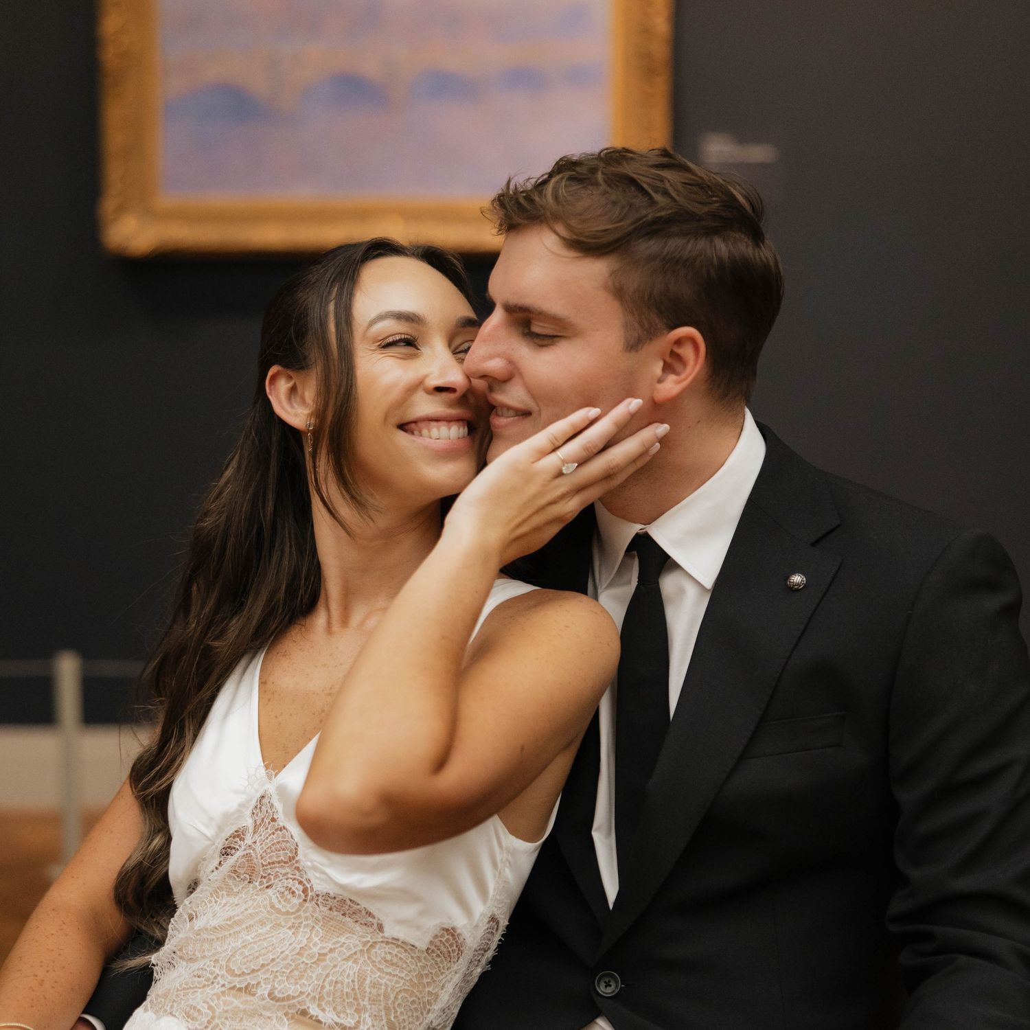 Happy couple sharing a romantic moment in an art gallery, woman showing engagement ring, man in black suit.