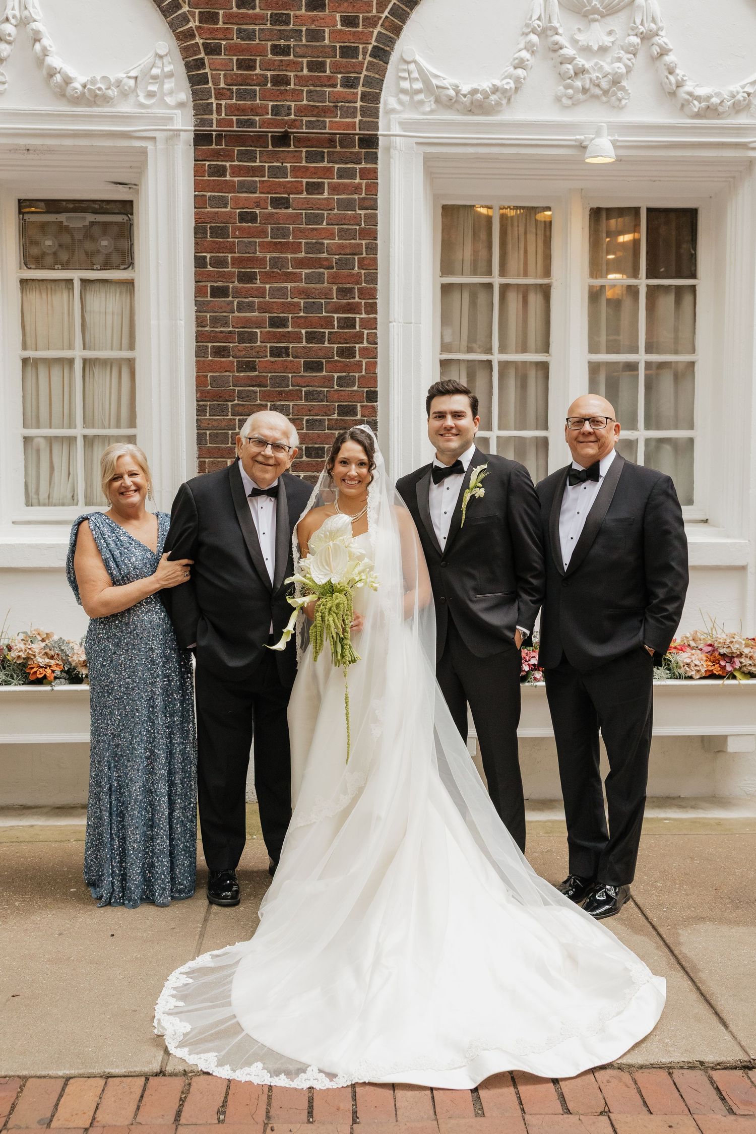 Bride and groom pose with family in formal attire outside brick building, bride wearing white gown with long train.