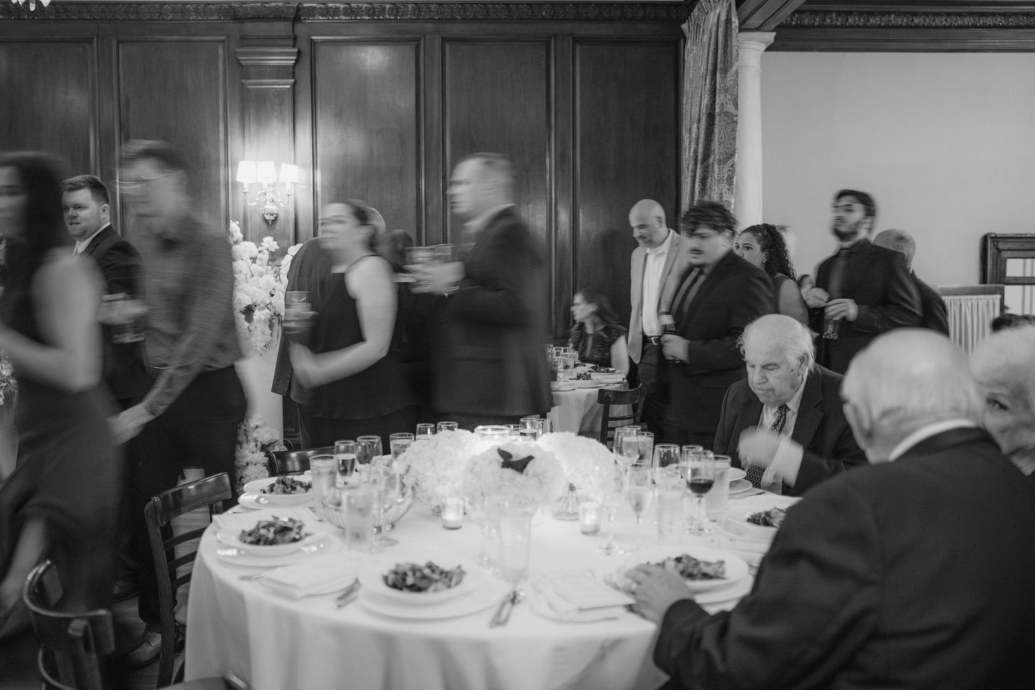 Black and white photo of elegantly dressed guests mingling and seated at a formal dinner reception in a wood-paneled ballroom.