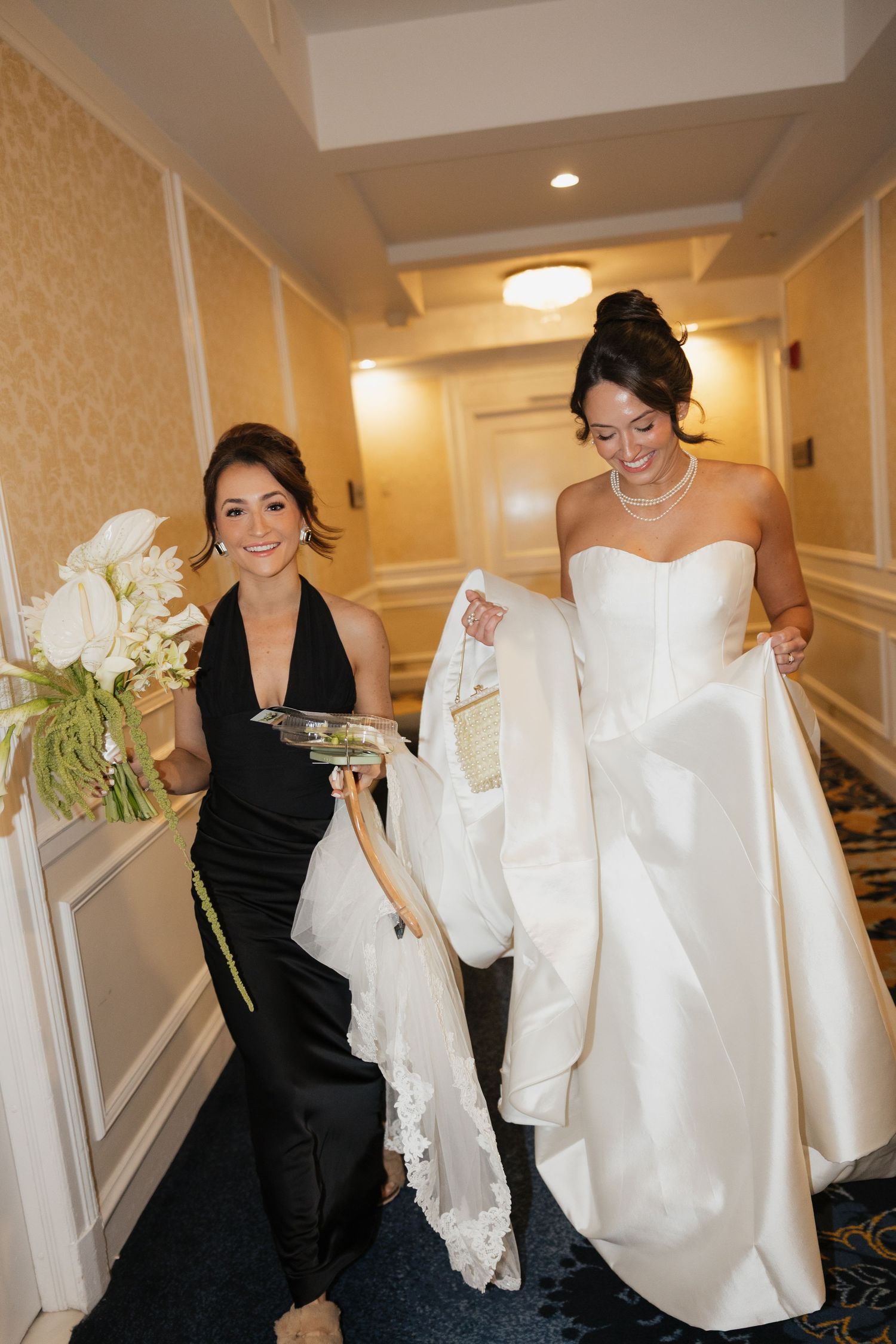 Bride in strapless white gown walks hotel hallway as bridesmaid in black dress carries bouquet and train.