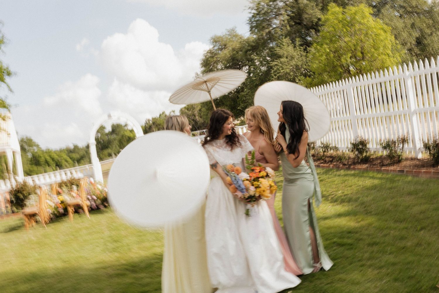 Bride and bridesmaids in colorful gowns holding white parasols in a sunny garden with white picket fence.