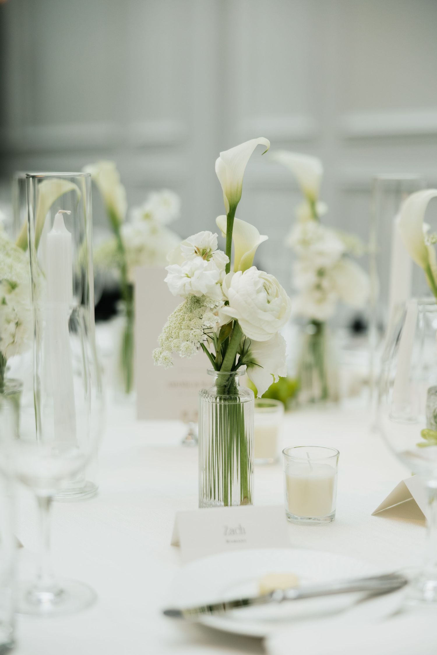 Elegant wedding reception table with white calla lily floral centerpiece, crystal glassware, and candles on white linen.