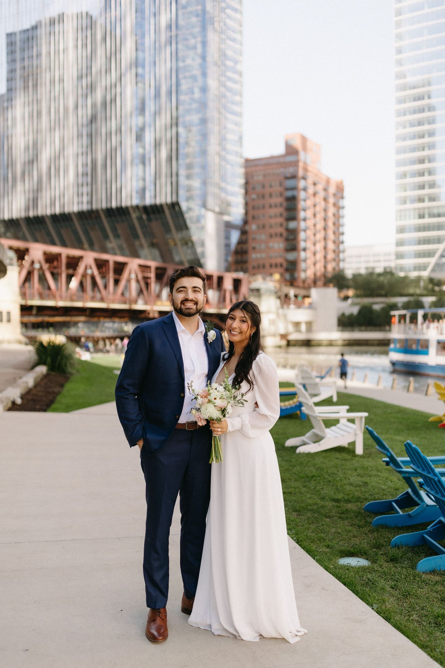 A couple in wedding attire shares a joyful moment together on a city rooftop with blue lounge chairs and skyscrapers behind them.