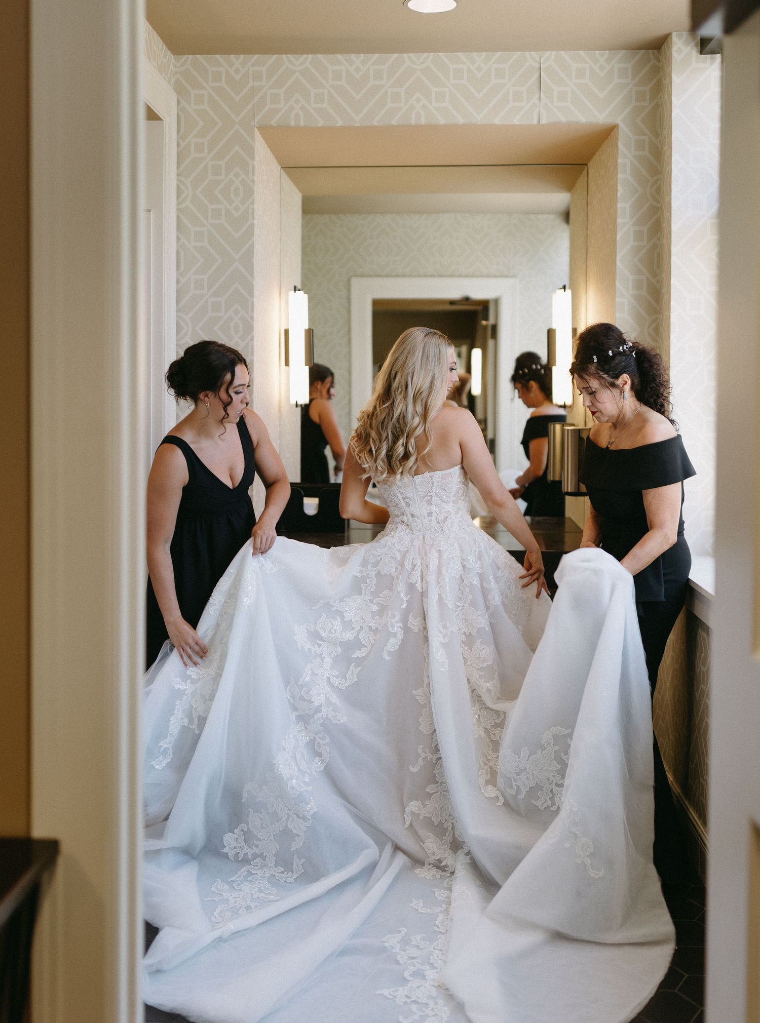 Bridesmaids in black dresses help adjust wedding gowns in an elegant hallway.