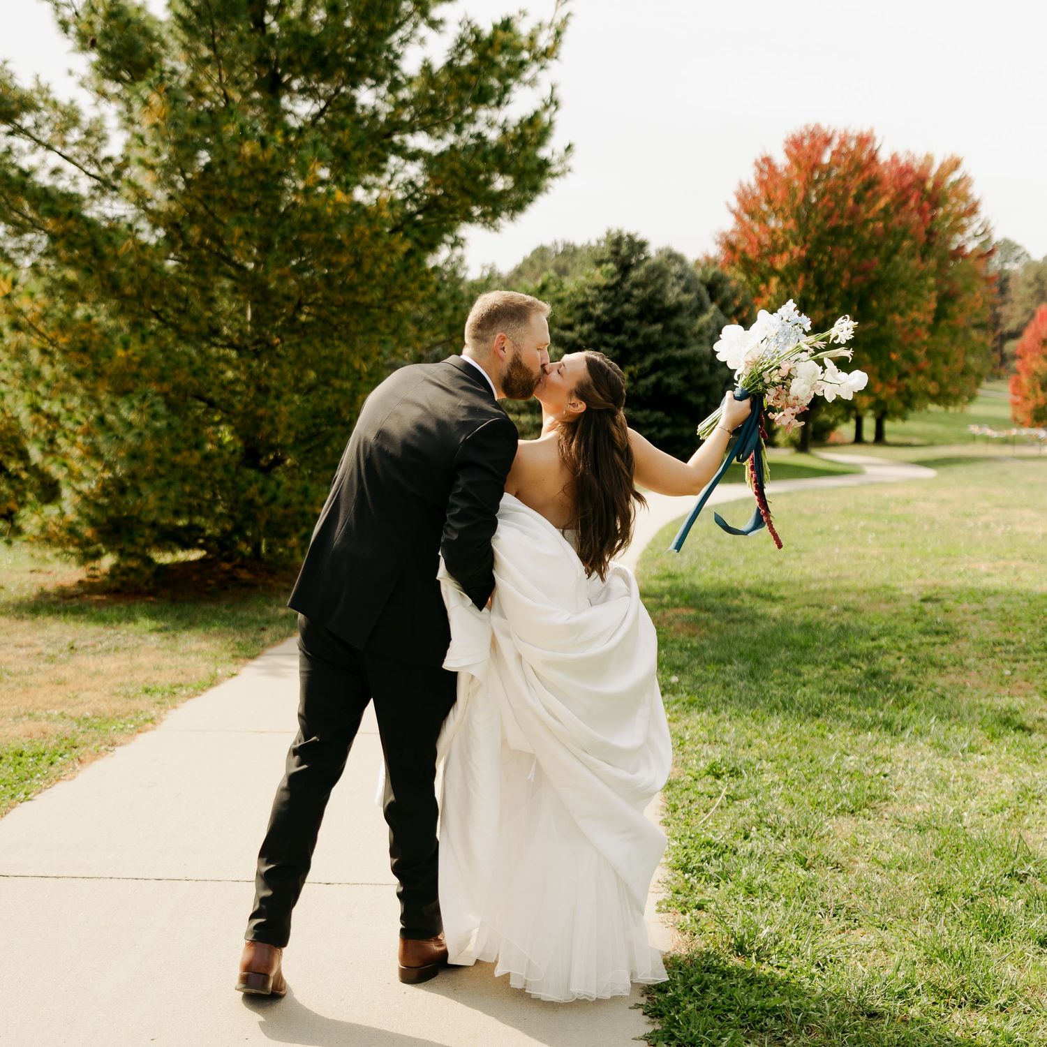 Bride in a white gown holding a bouquet kisses her groom in a dark suit on a sunny park path with autumn trees.