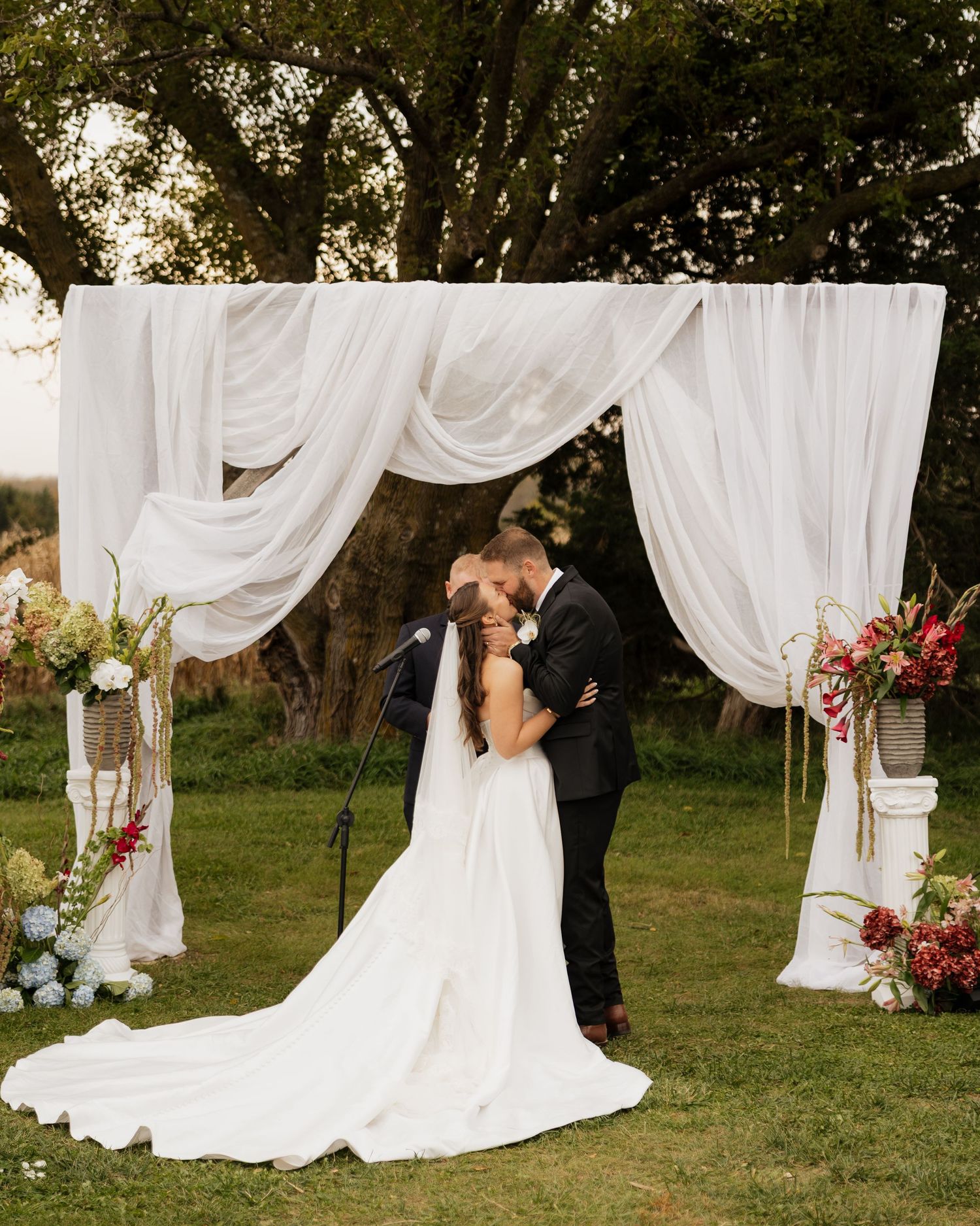 Bride and groom share a kiss beneath a draped white fabric arch with colorful flower arrangements at an outdoor wedding ceremony.