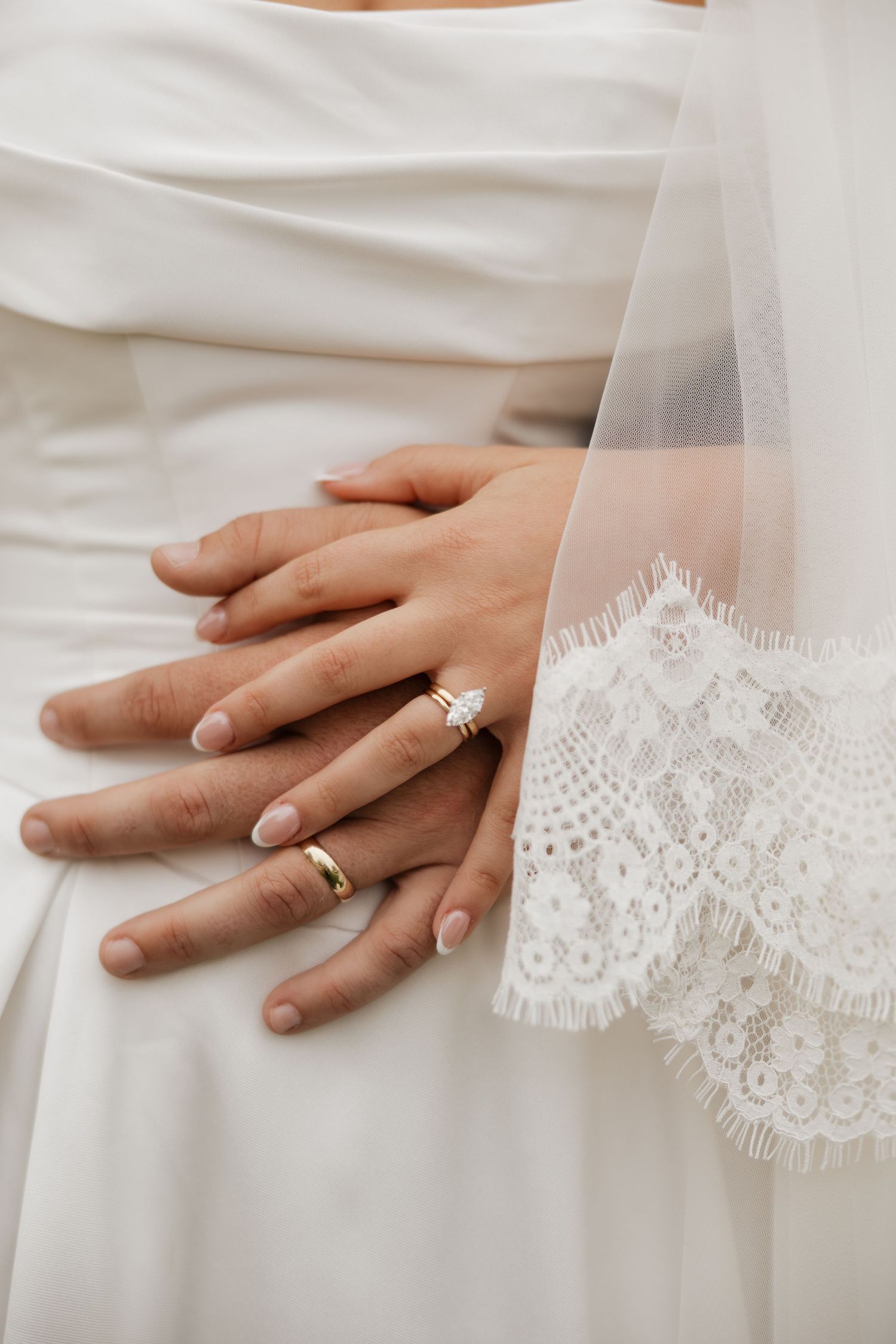 Bride and groom's hands layered on a white wedding gown, showing a marquise diamond engagement ring, gold band and lace veil edge.
