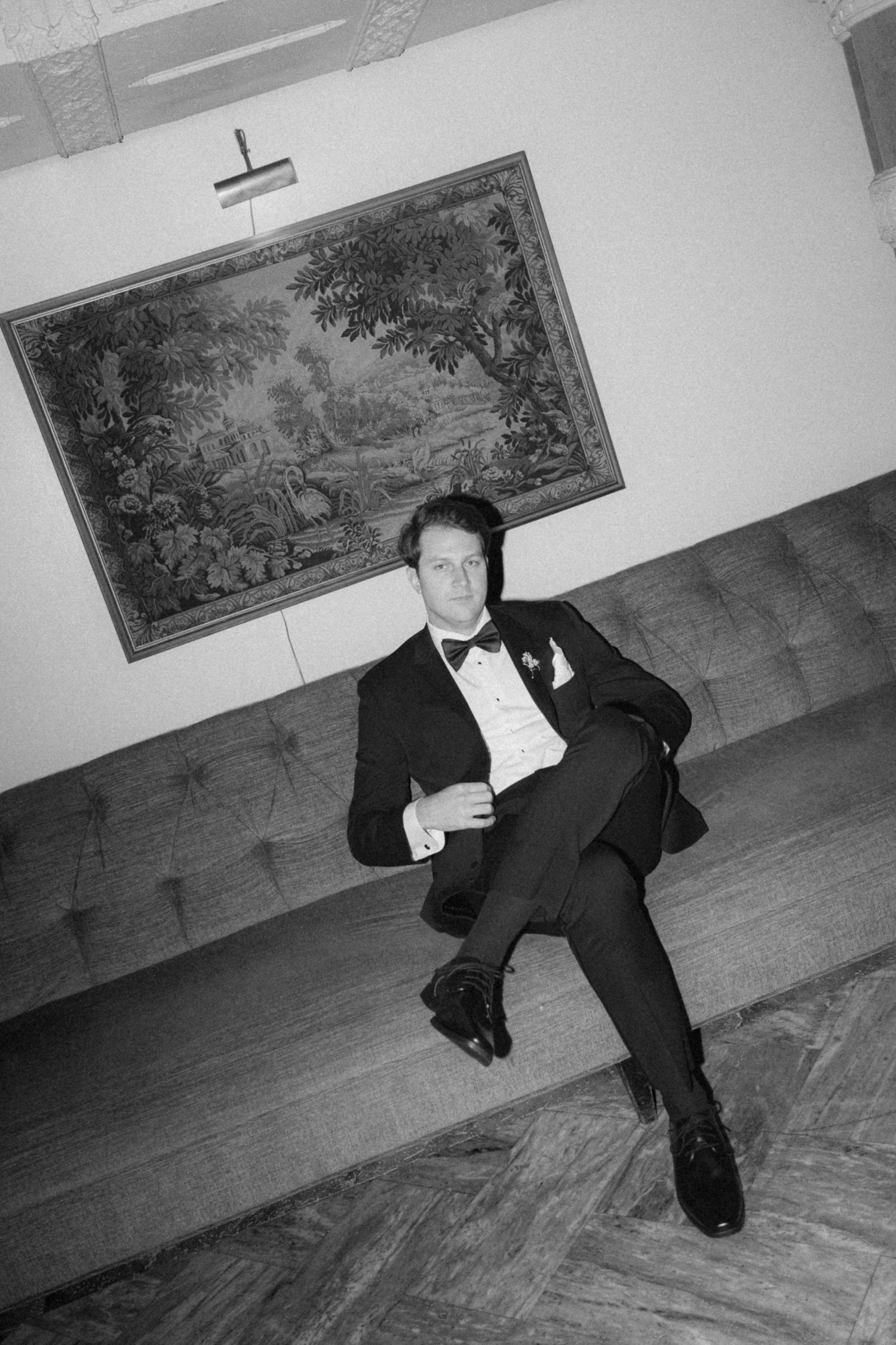 Black and white photo of a man in a tuxedo and bow tie reclining on a tufted sofa beneath a framed woodland tapestry.