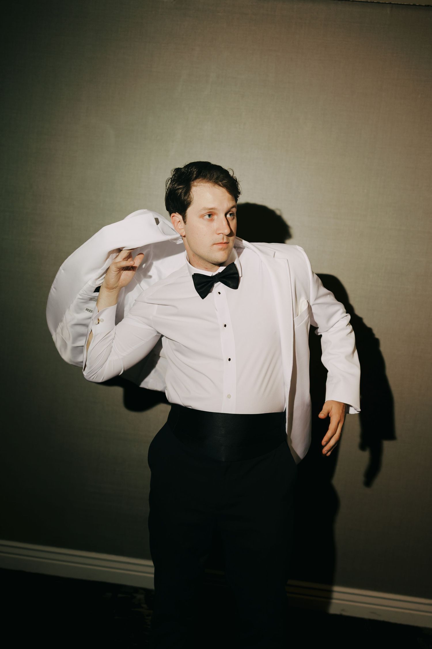 Groom in black bow tie and cummerbund holds a white tuxedo jacket over his shoulder while posing against a dark wall.