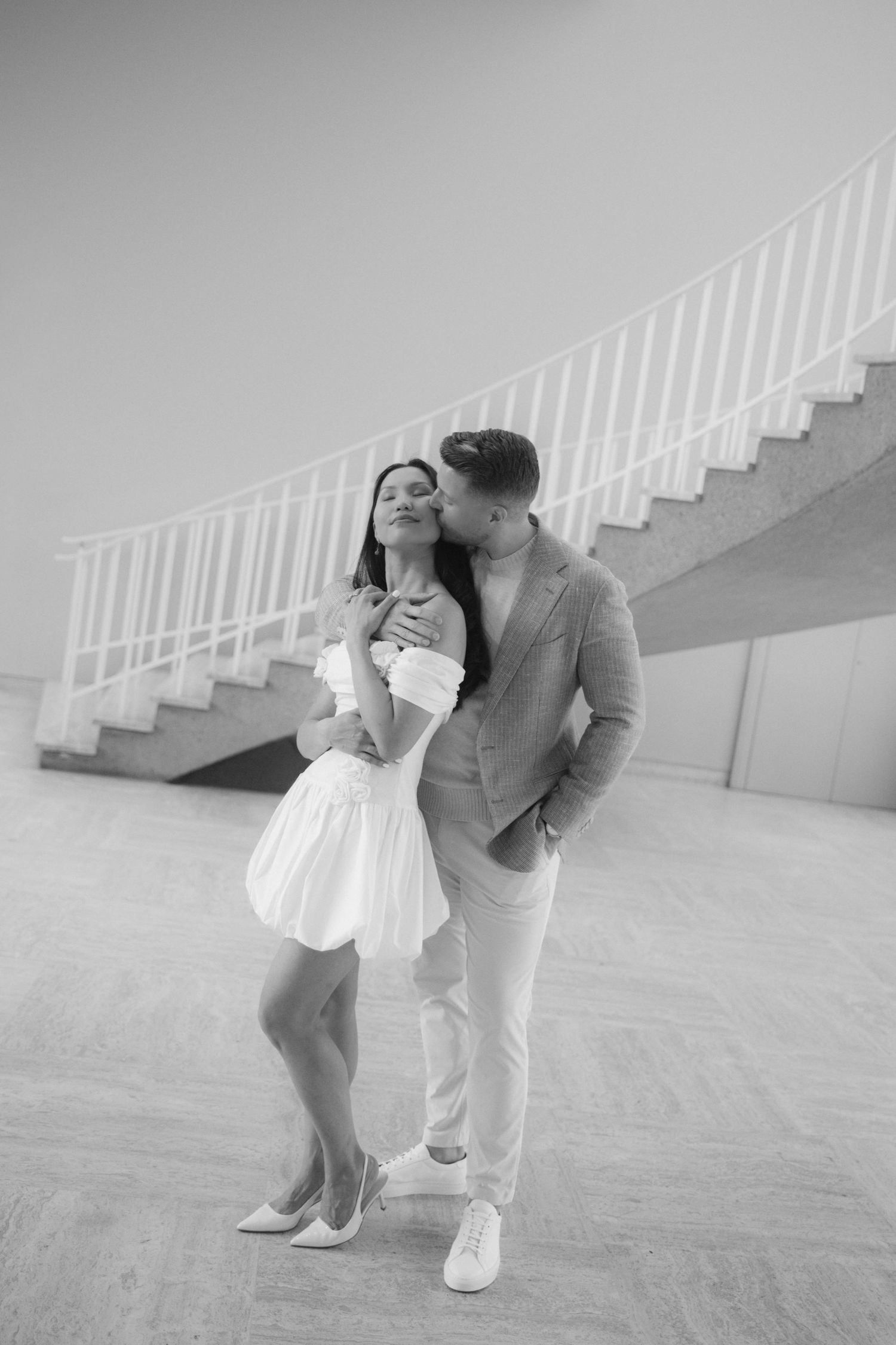 Black and white photo of a romantic couple embracing near a staircase, man kissing woman in white dress.
