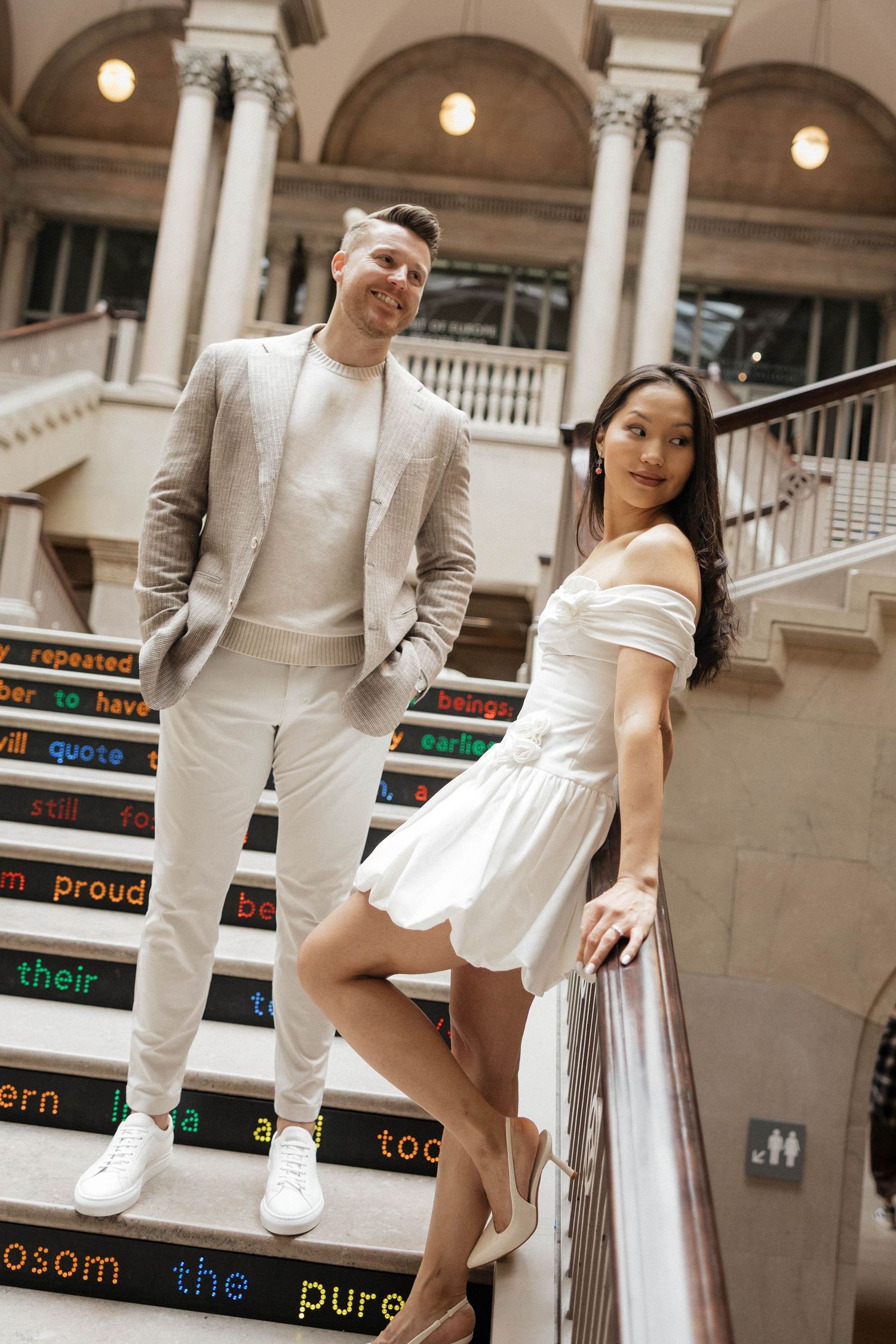 Couple posing on colorful text-decorated stairs in elegant white outfits inside a grand building with columns.