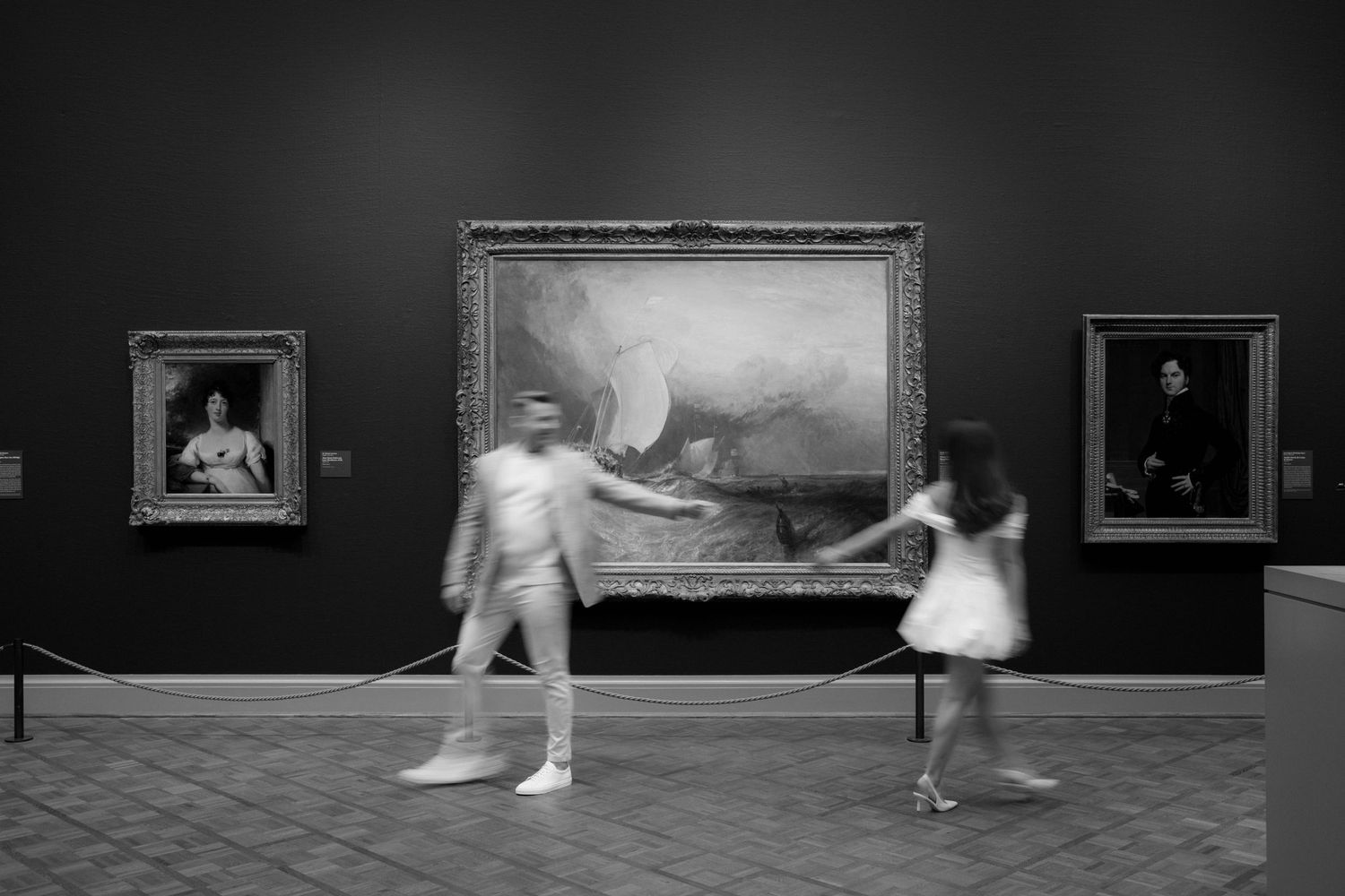 Black and white photo of blurred visitors walking past large paintings in a dimly lit art museum gallery.