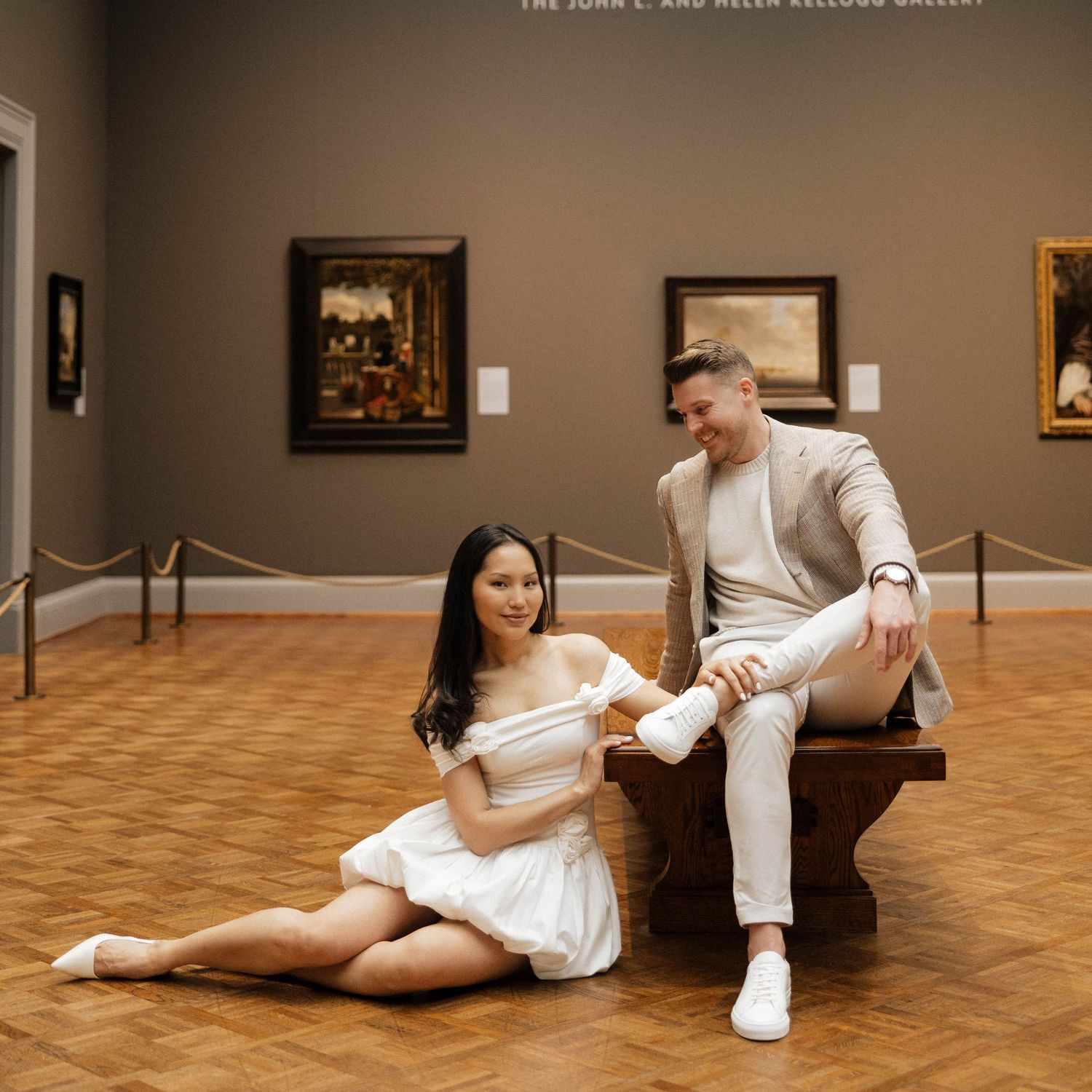 Couple posing in art museum gallery, woman in white dress sitting on floor, man in beige suit on bench.