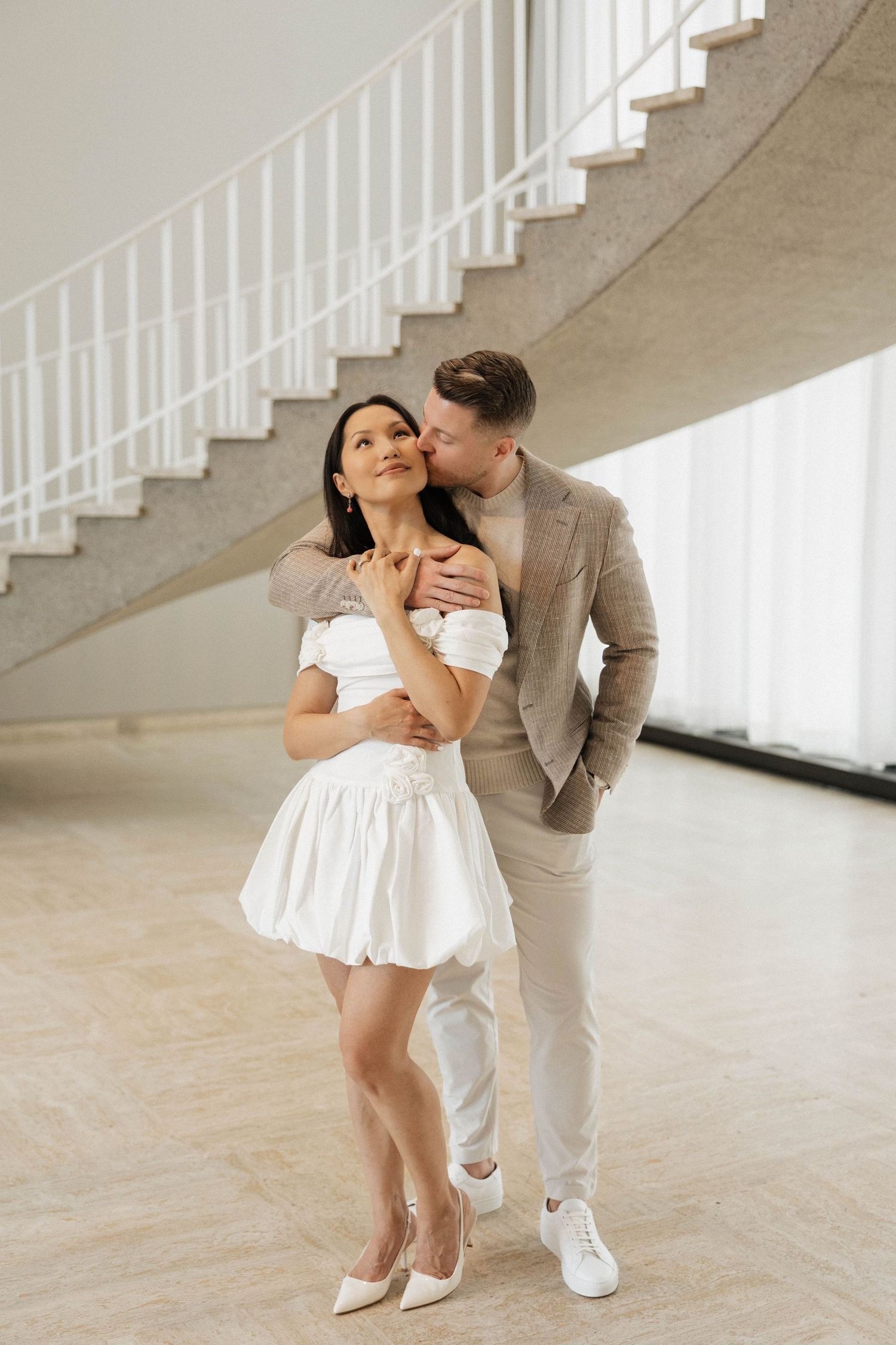 Elegant couple embracing near spiral staircase, woman in white mini dress and heels, man in beige suit kissing her cheek.