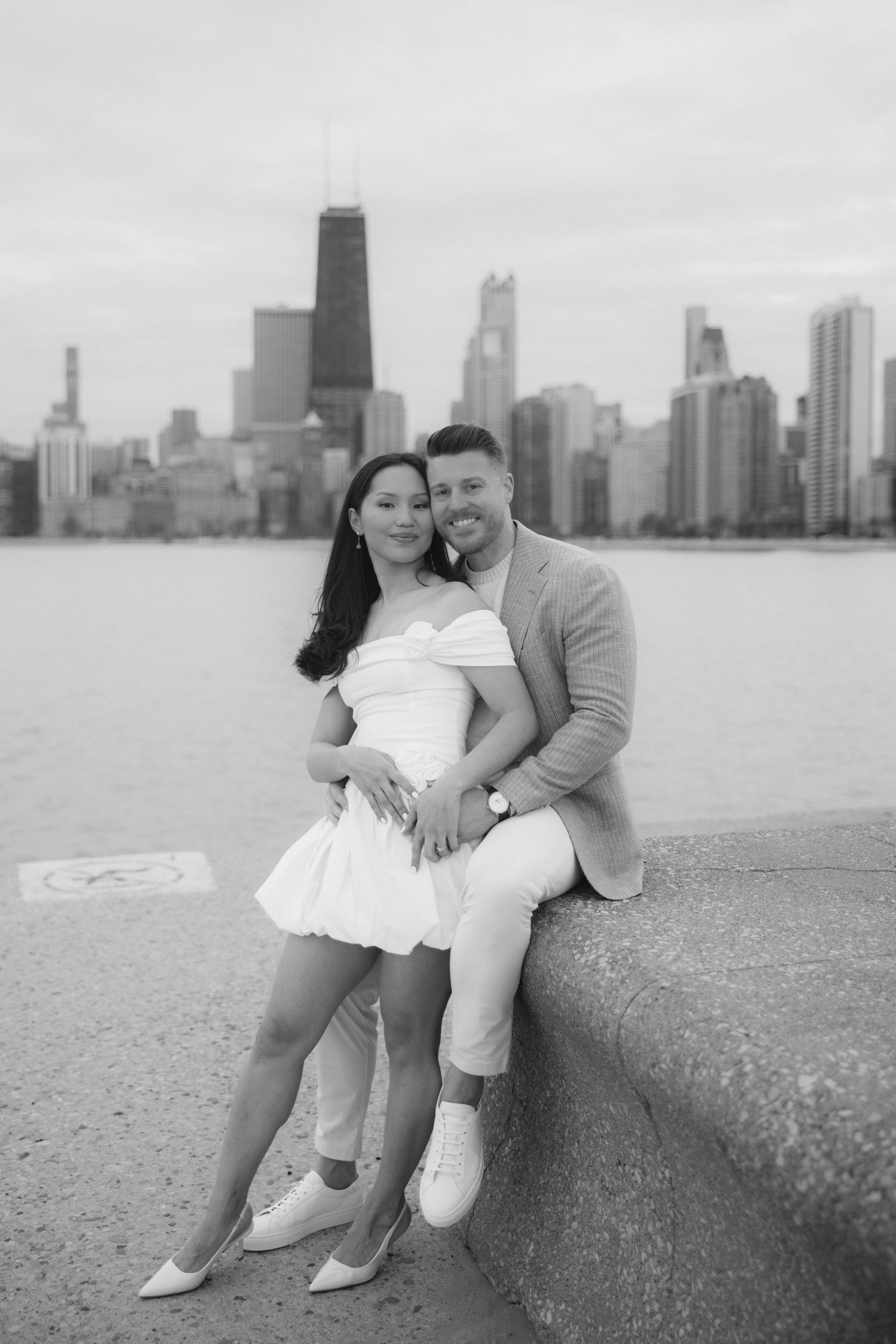 Couple poses by Chicago lakefront with city skyline in black and white engagement photo.