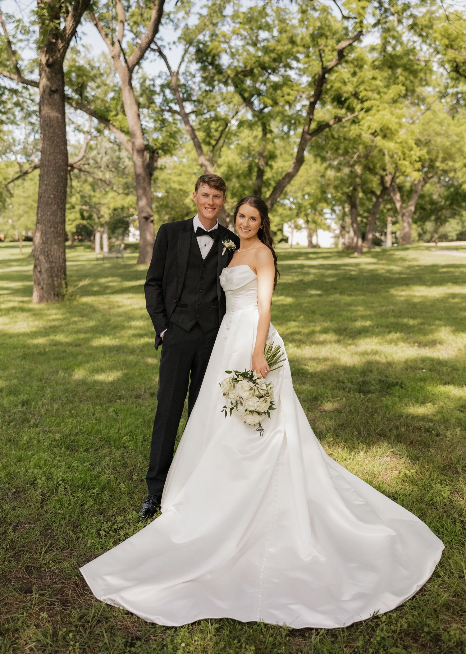 Bride in white strapless gown with train and groom in black tuxedo pose together in a lush green park setting.
