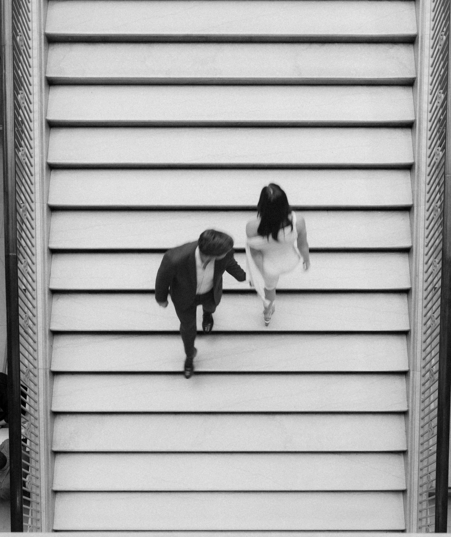 Black and white overhead photo of a man in a suit and a woman in a white dress walking down a wide staircase.