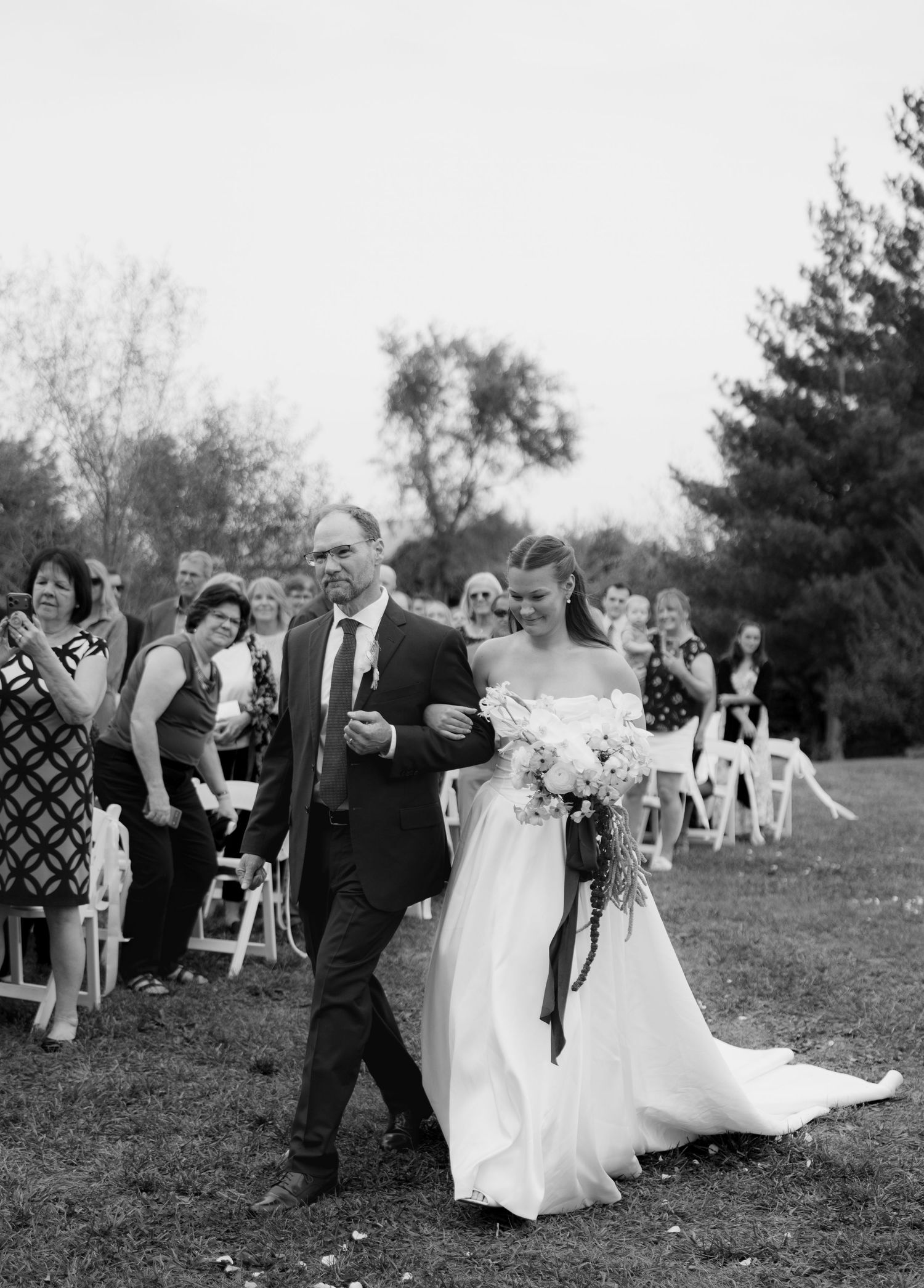 Black and white photo of a father walking his daughter, a bride in a strapless gown with a bouquet, down the outdoor wedding aisle.