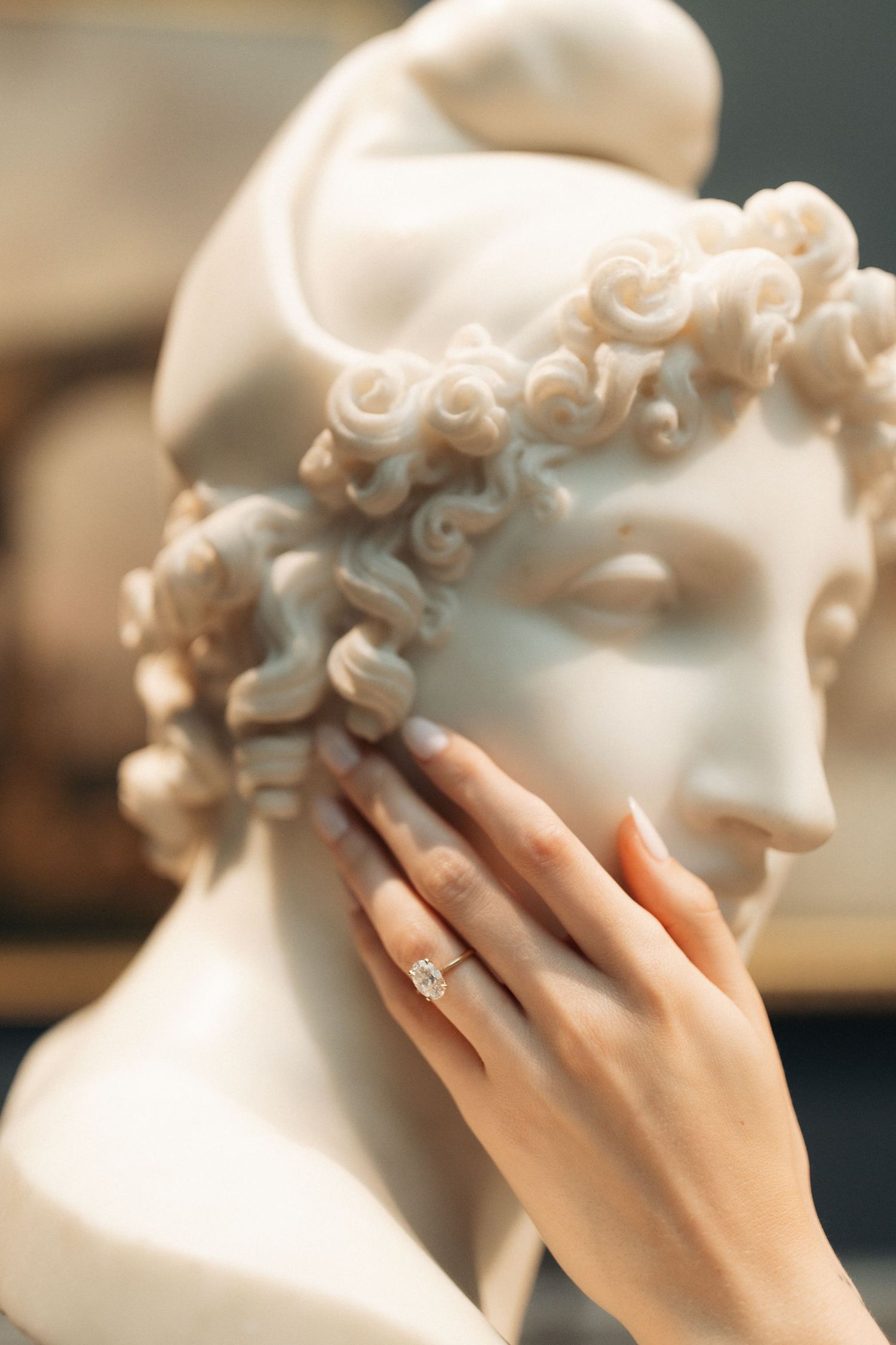 A hand wearing a diamond engagement ring gently touches a classical marble bust sculpture.
