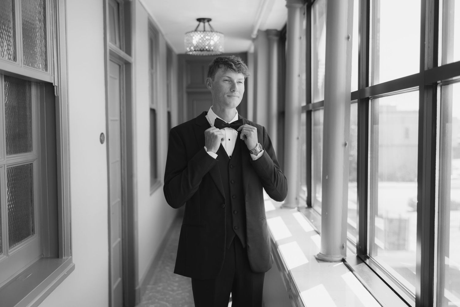 Groom in black tuxedo adjusting bow tie in elegant hallway, black and white wedding photography.