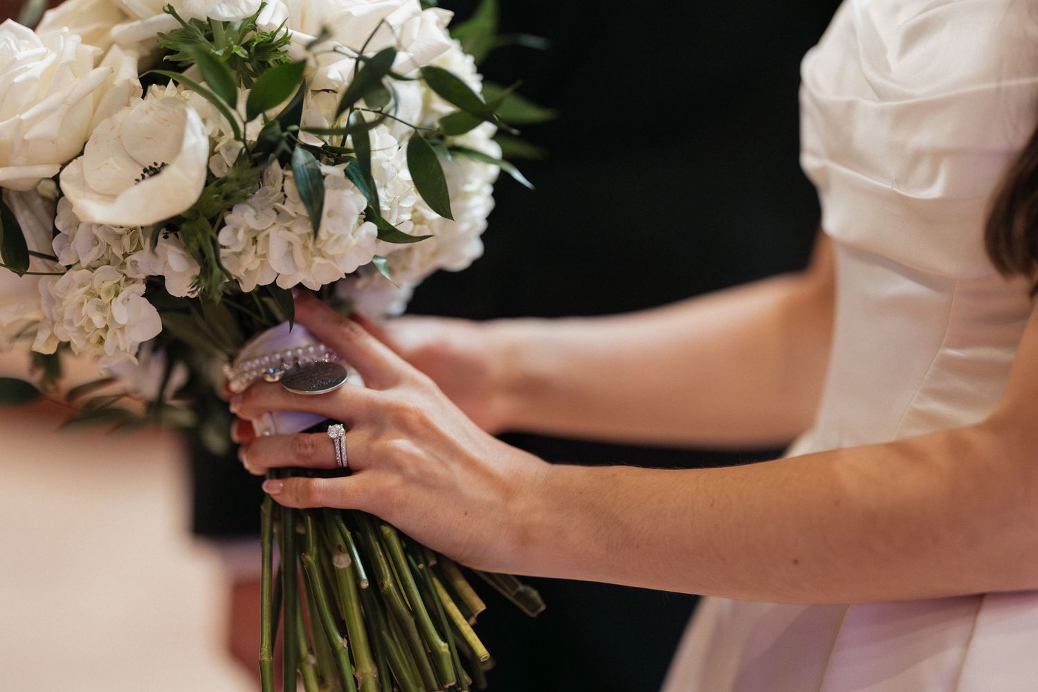 Bride holding a white floral wedding bouquet with roses and greenery, wearing a diamond ring and white dress.
