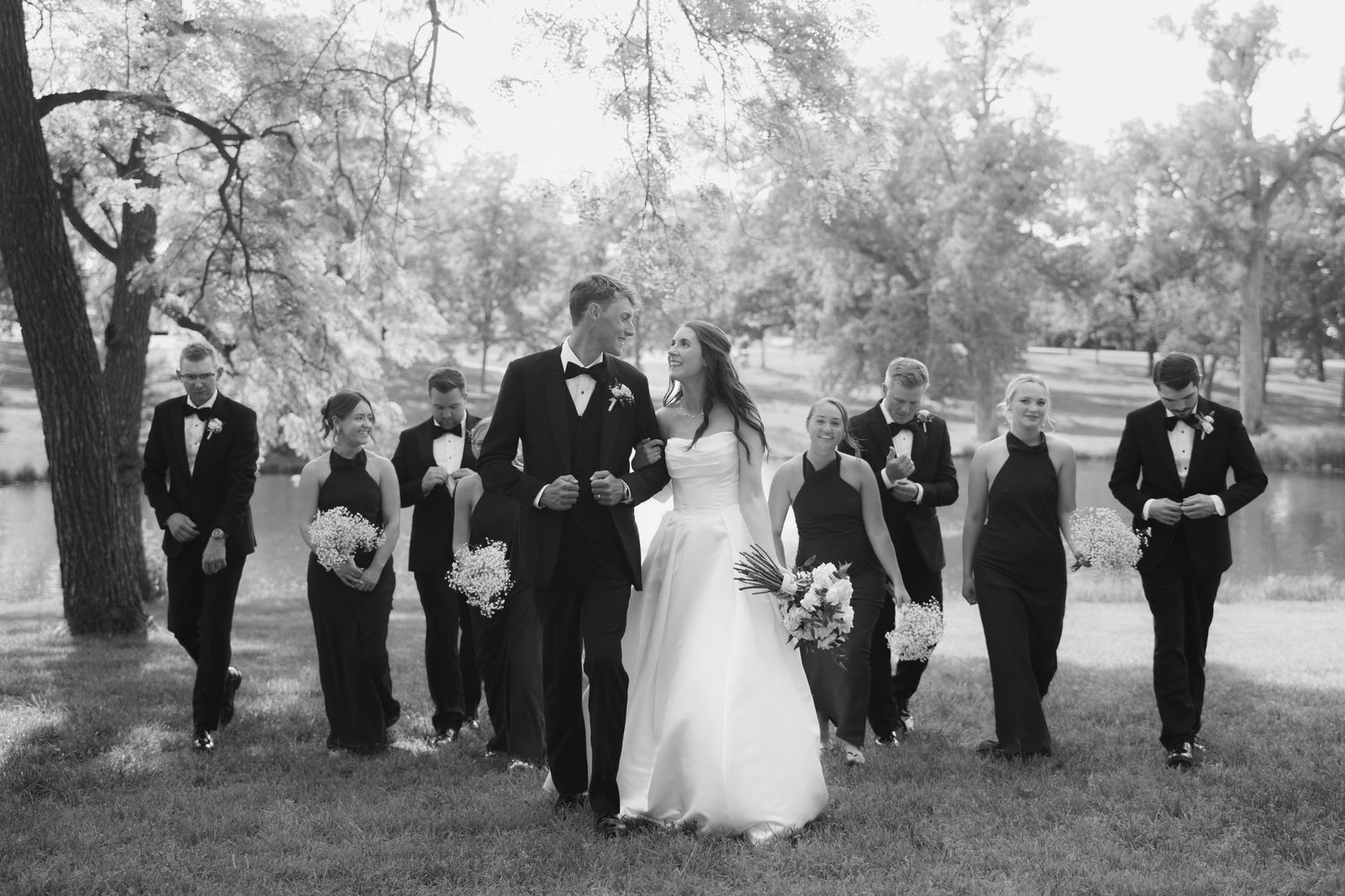 Black and white wedding party walking outdoors near trees and pond, bride and groom kissing in center.