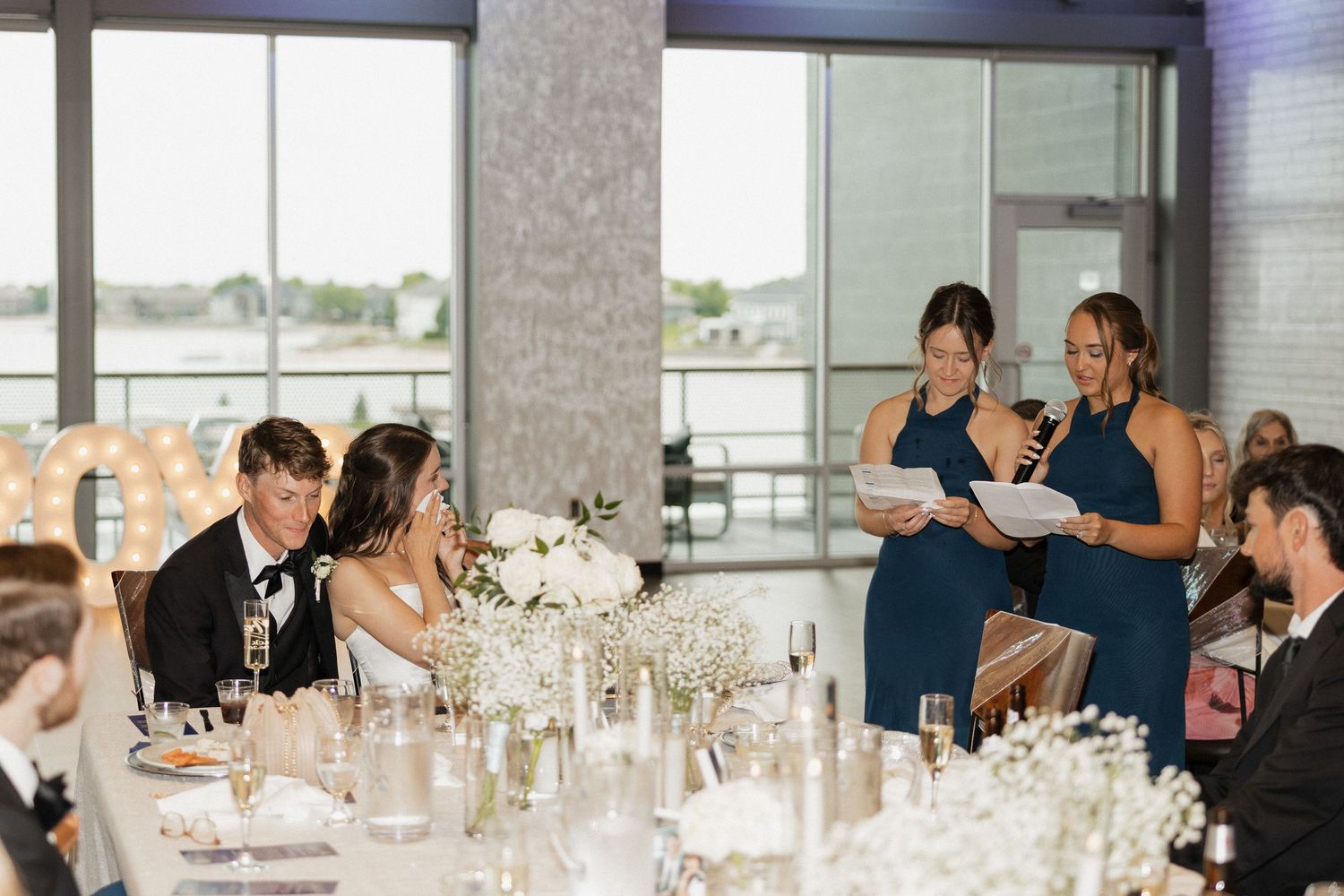 Two bridesmaids in blue dresses give a toast at an elegant waterfront wedding reception with white floral centerpieces.