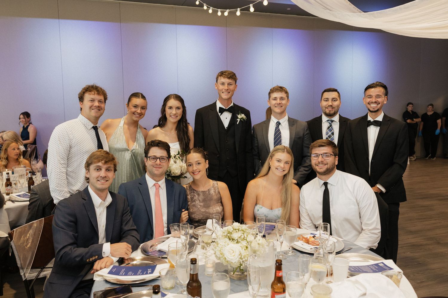 Group of elegantly dressed guests smiling together at a wedding reception dinner table in a decorated banquet hall.