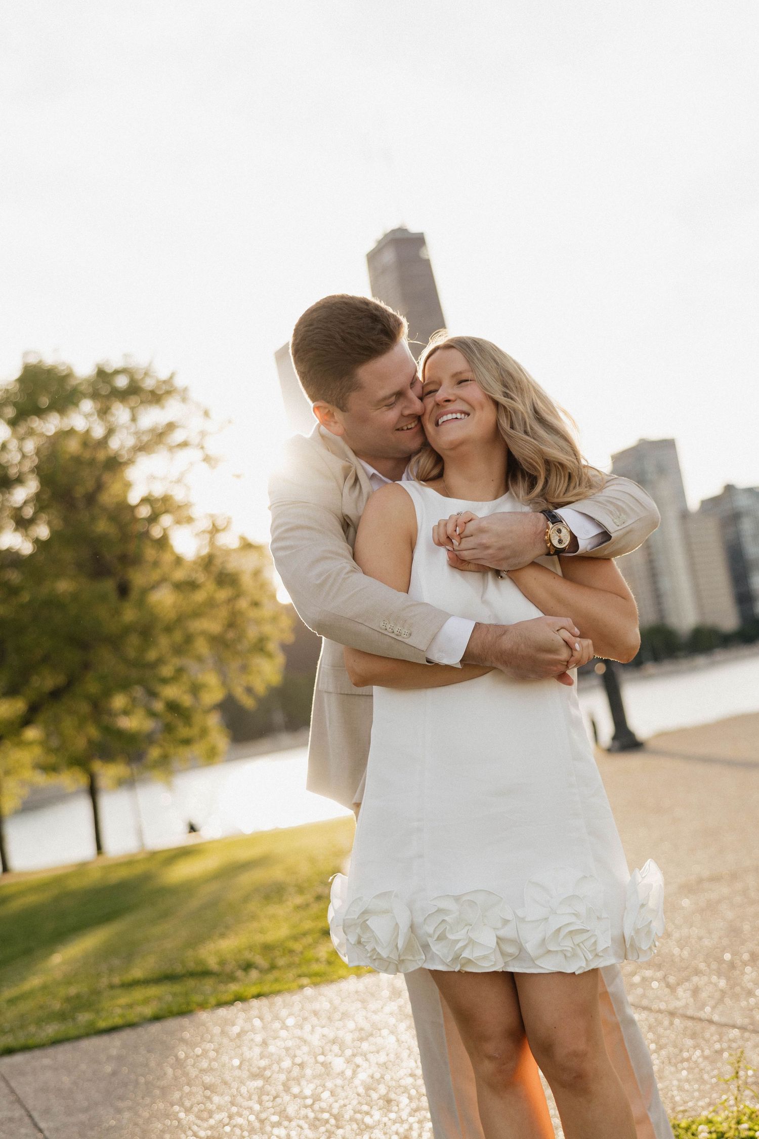 Man embracing woman in white dress by a lakeside park with city skyline backdrop.