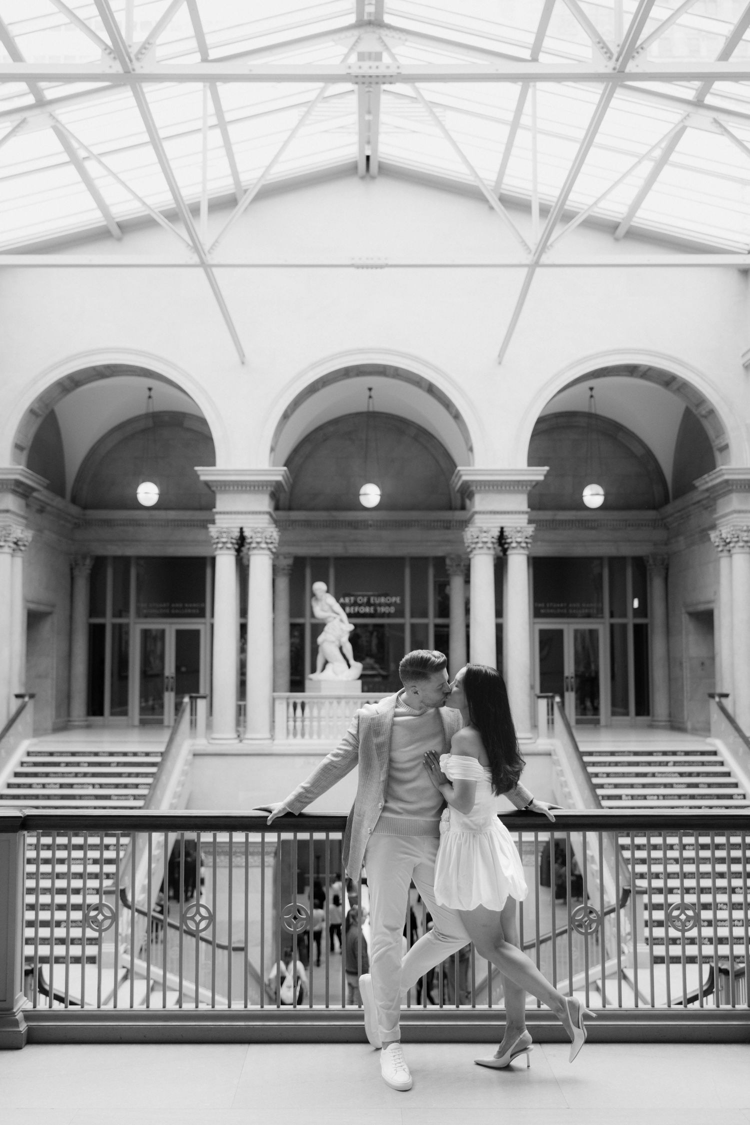 Black and white photo of a couple walking in an elegant museum atrium with arched columns and a classical sculpture.