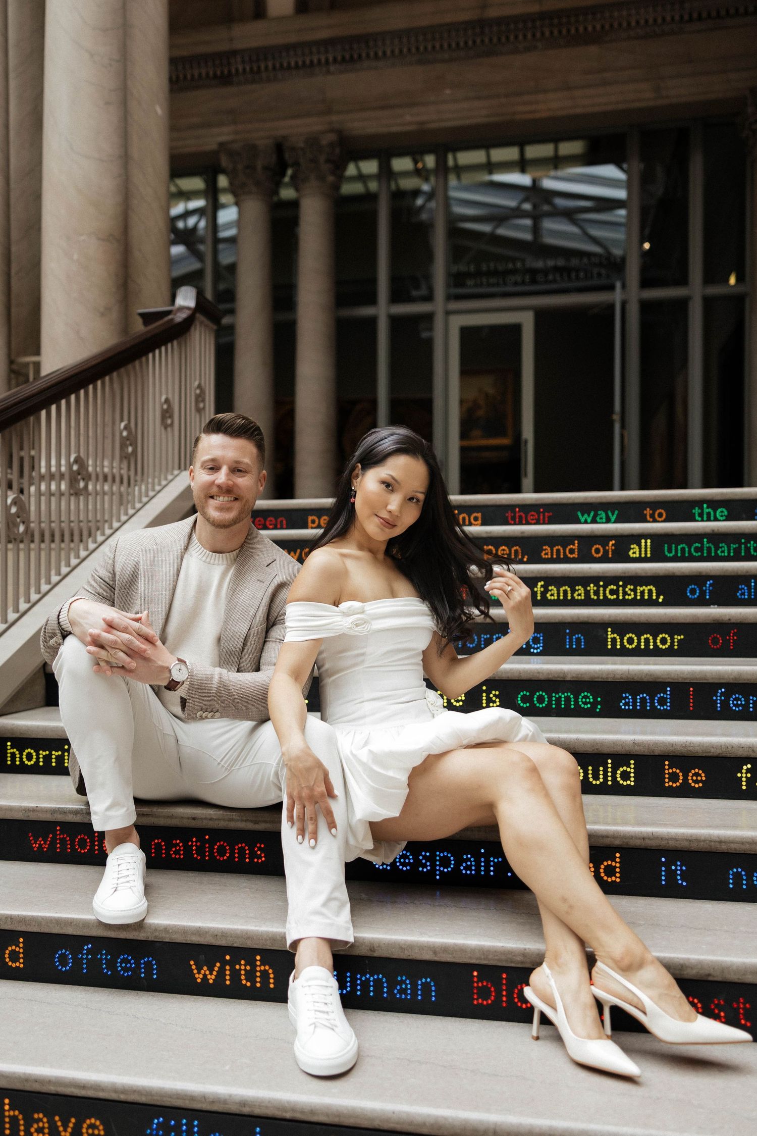 Couple in white outfits sitting on colorful text-inscribed steps outside a grand building, smiling at camera.