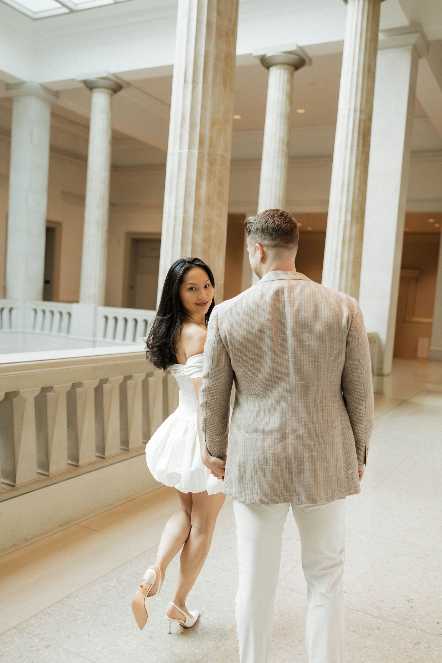 Couple holding hands in elegant museum hall with white columns, woman in white dress looks back smiling at man in beige blazer.