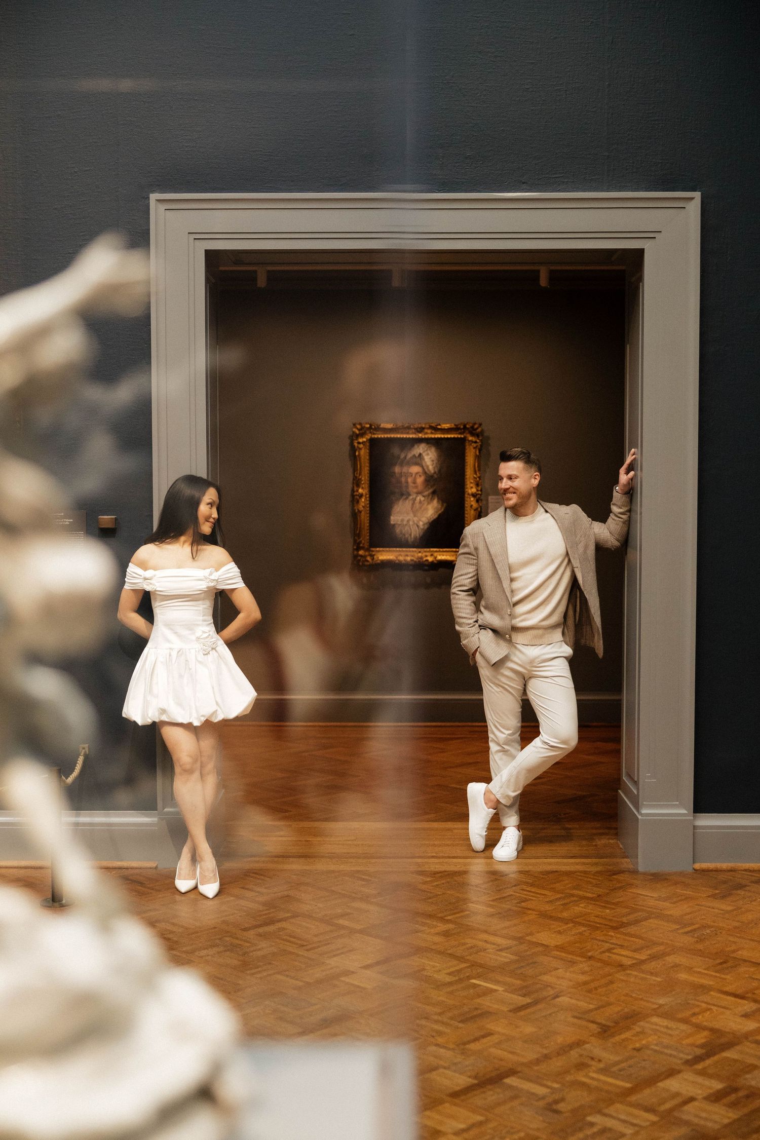 Couple posing in elegant art museum gallery with classic portrait painting and hardwood floors.