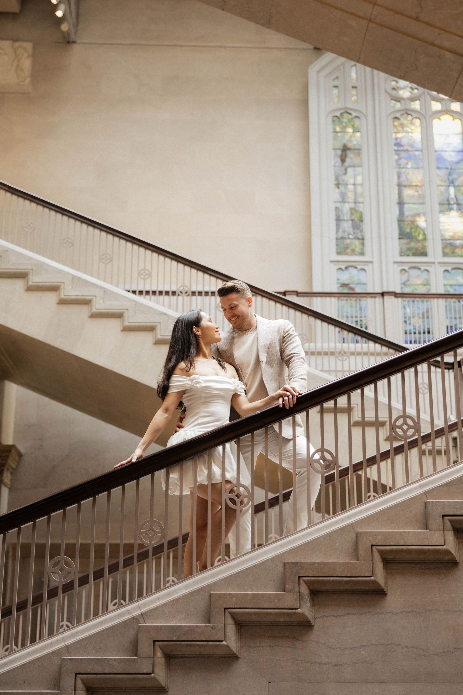 Elegant couple shares romantic moment on grand staircase with stained glass window backdrop.