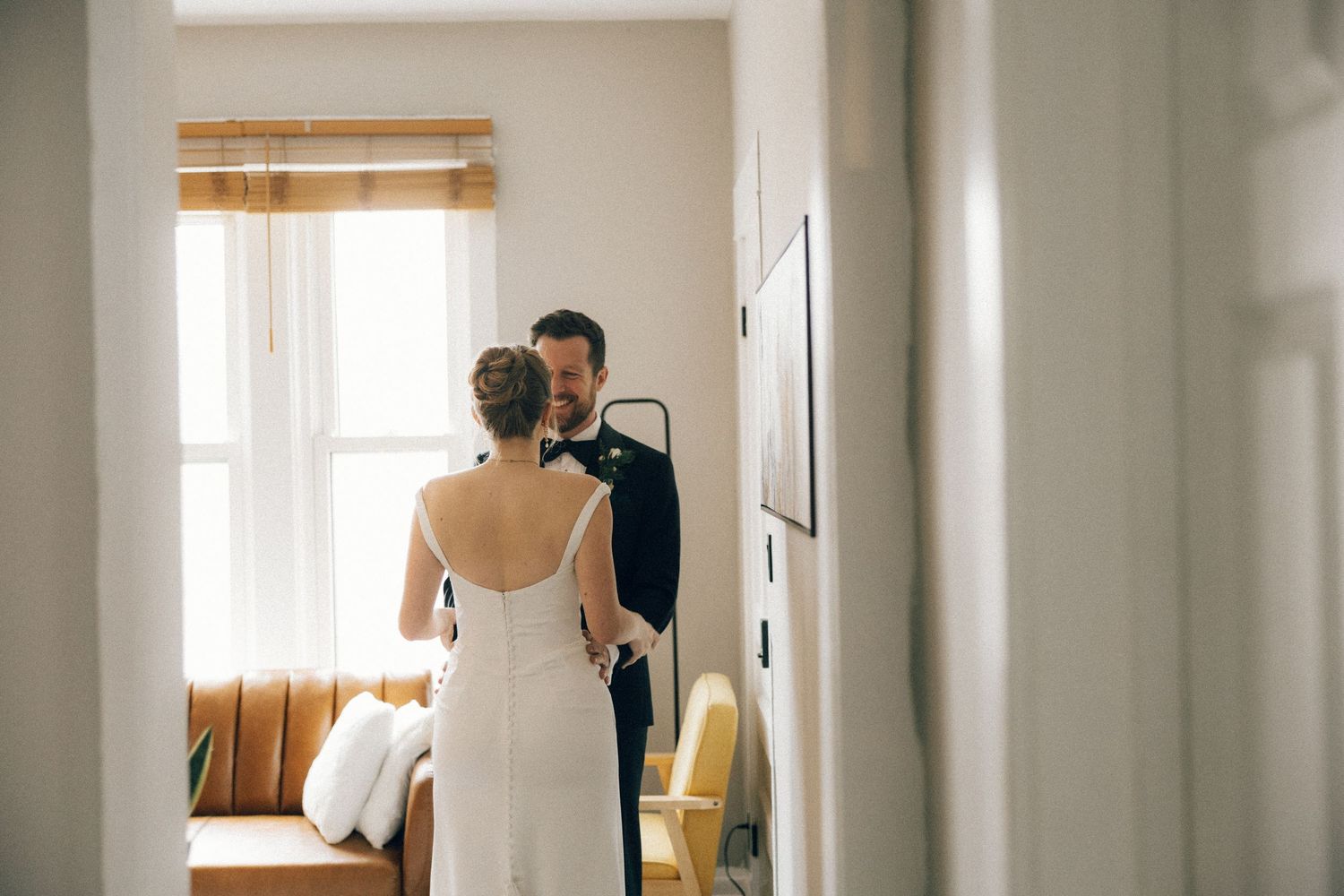 A bride in a white backless wedding dress stands in a bright sunlit room near bamboo window shades.